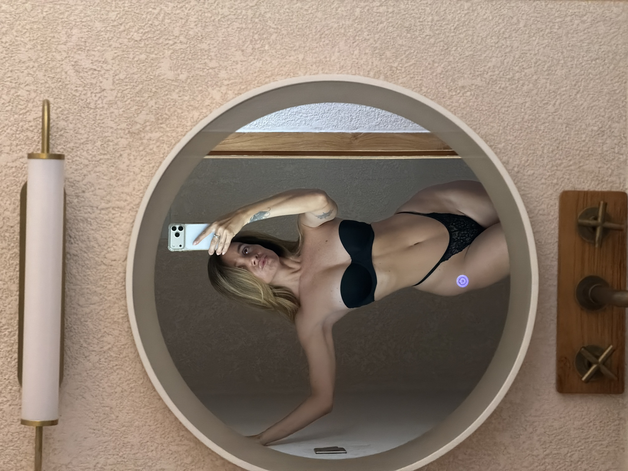 A woman in her 20s or 30s, with shoulder-length blonde hair, is taking a mirror selfie. She is wearing a black strapless bra and black lace bikini bottoms. She is standing with her right arm extended, holding a smartphone to take the photo. Her left arm is bent, with her hand resting on the edge of the mirror. Her body is angled towards the camera, and she is making a kissy face. The mirror is round and framed in a light color. The background appears to be a bathroom with textured walls and a light fixture above the mirror. The photo was taken in a domestic setting in the city of Mengwi, Indonesia.