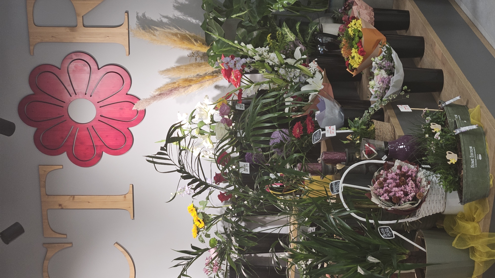 The image shows a flower shop display, likely in Prešov, Slovakia, given the presence of Slovak currency on some labels. The backdrop is a light gray wall with large wooden letters spelling out "FLOWER" and a red-pink wooden flower cutout. The foreground is filled with a variety of floral arrangements and potted plants displayed on tiered shelving.
On the top shelf, a large red-pink flower cutout is prominently displayed on the wall, flanked by wooden letters. Below this, a collection of plants and bouquets are arranged. To the left, there are sunflowers, roses, and green leafy plants in various containers. In the center, there are tall palm-like plants, red roses, lilies, and other colorful flowers. To the right, there are more lilies, red anthuriums, and tall dried grass plumes.
The middle shelf features more potted plants and smaller bouquets. On the left, a large potted Yucca plant with a price tag of "44,50€" is visible. Next to it, a white wicker basket holds a bouquet of dried pink flowers and pinecones. A purple vase sits nearby. In the center and right of this shelf, there are more floral arrangements in vases and pots, including some with purple and pink flowers. Smaller plants with decorative watering can figures are in the front row, with one pot labeled "HOME EDITION VINTAGE CONTAINER SINCE 1992."
The overall impression is a well-stocked and visually appealing flower shop display, with a focus on diverse floral offerings. The lighting appears to be artificial, likely from overhead shop lights, creating a well-lit but somewhat uniform illumination of the scene.