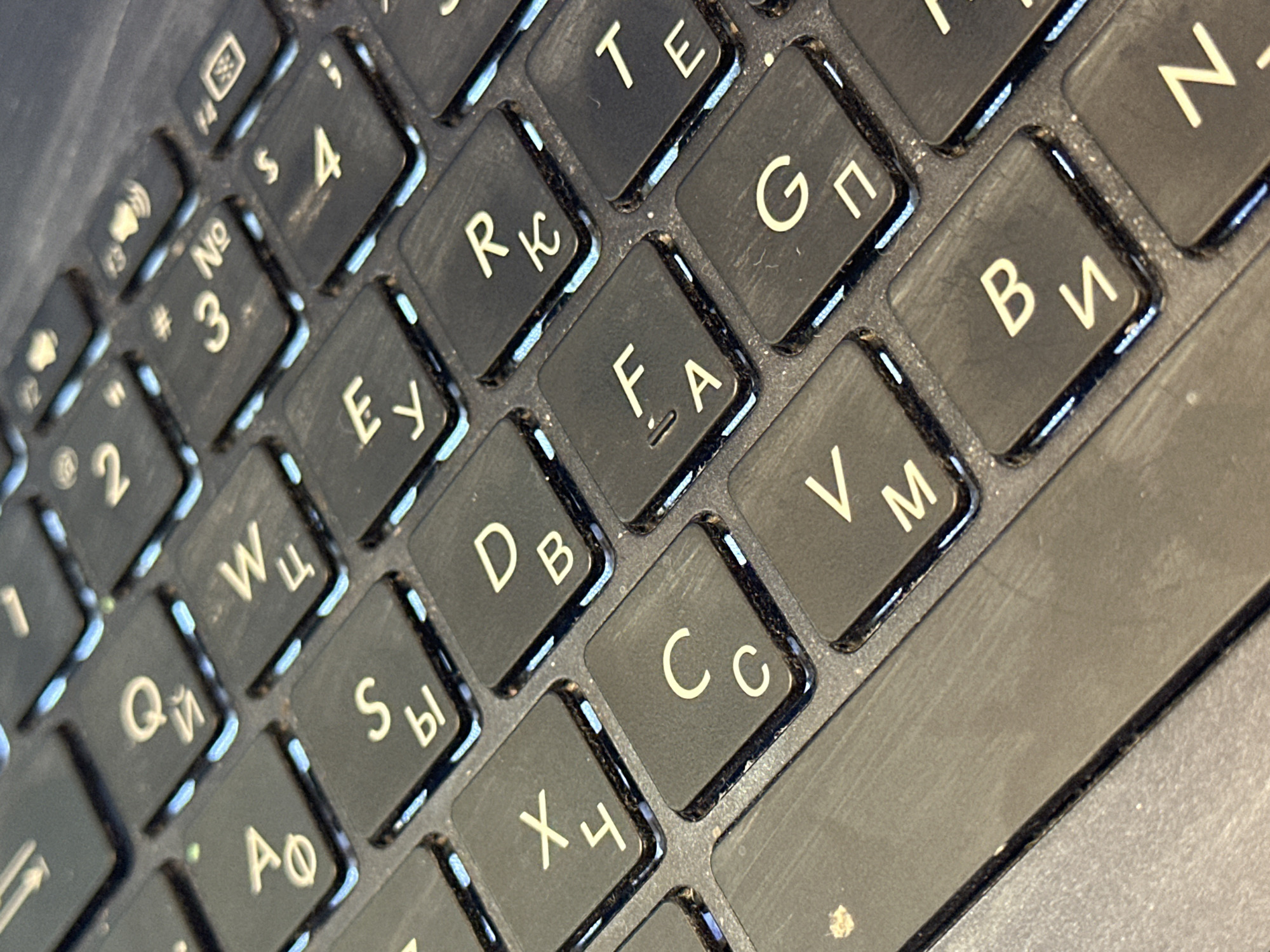 The image shows a close-up, high-angle view of a black keyboard with illuminated white Cyrillic letters and symbols. The keys are slightly dusty, and the lighting suggests they are backlit. Some keys visible include "Q" (represented by a Cyrillic letter that looks like a "й" or "i"), "W" (represented by a Cyrillic "ц"), "S" (represented by a Cyrillic "ы"), "A" (represented by a Cyrillic "ф"), "E" (represented by a Cyrillic "у"), "D" (represented by a Cyrillic "в"), "R" (represented by a Cyrillic "к"), "F" (represented by a Cyrillic "а"), "T" (represented by a Cyrillic "е"), "C" (represented by a Cyrillic "с"), "G" (represented by a Cyrillic "п"), "V" (represented by a Cyrillic "м"), "B" (represented by a Cyrillic "и"), and "N" (represented by a Cyrillic "т"). There are also number keys with symbols, such as "3" with a "#" symbol and "No". The image does not contain any information that would allow for determining its location in Nevinnomyssk, Russia, or any other specific city. There are no people, actions, or interactions depicted. The environment is limited to the keyboard itself, which appears to be part of a laptop.