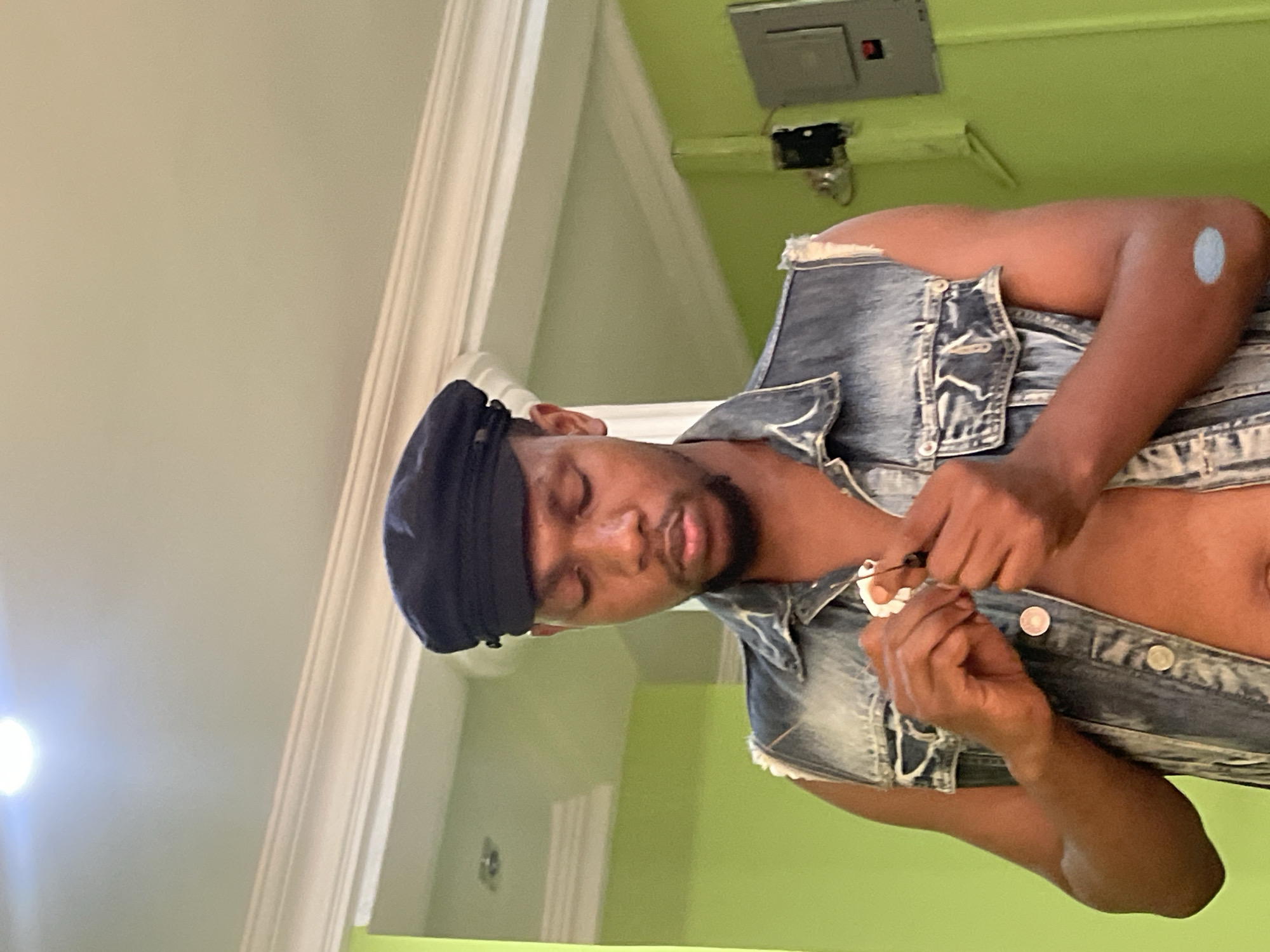 A man with dark hair and a black head wrap is fastening a denim vest with silver buttons. He is indoors. The wall is two-toned: a light cream color on the left and a bright green on the right. There is a silver electrical box on the upper right of the wall. The image does not have enough information to determine the location in the city of Abuja, Nigeria.