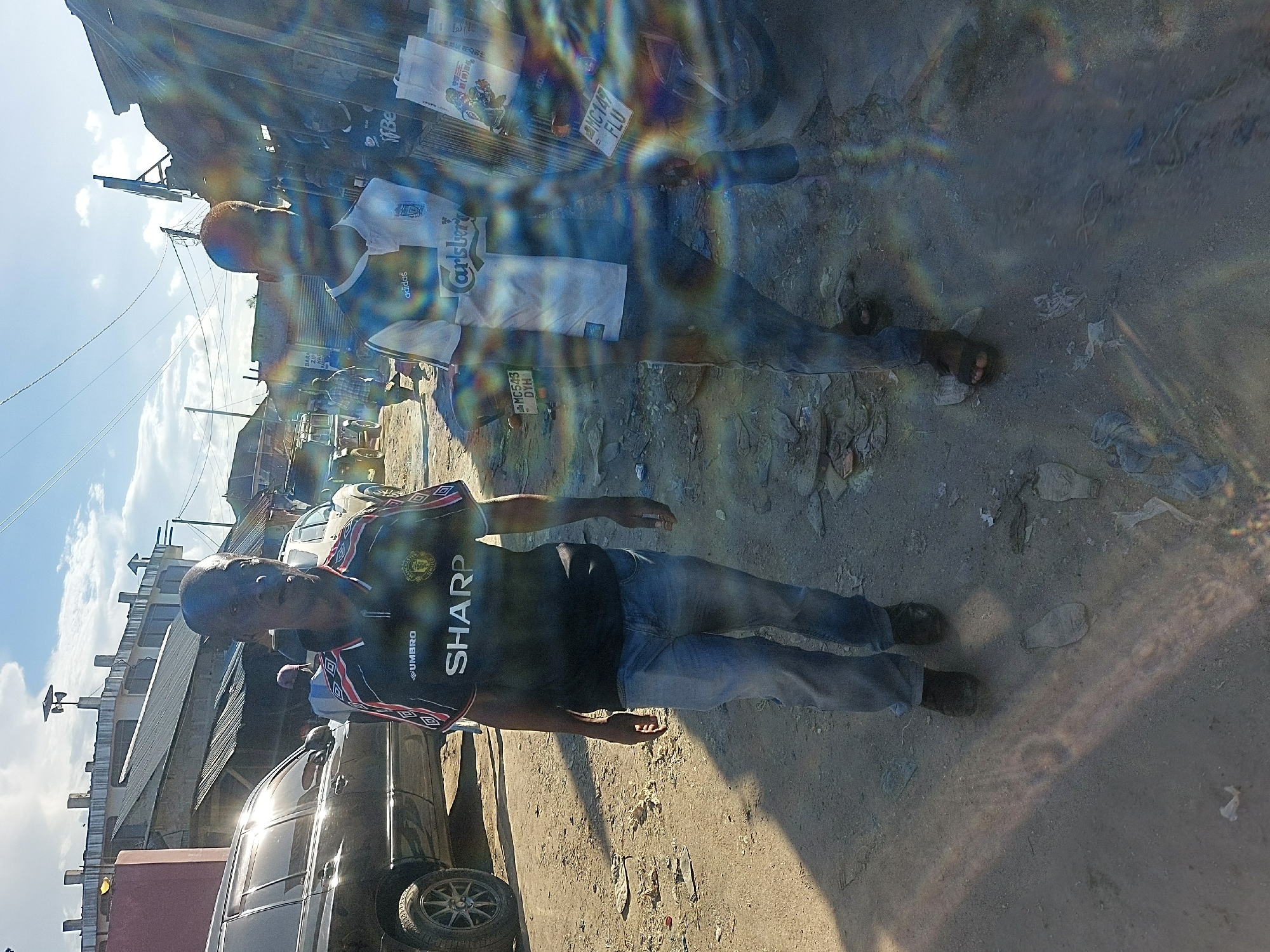 The image shows an urban scene in Dar es-Salaam, Tanzania, with two men standing outdoors on a dirt and gravel surface. The first man, closer to the camera, is wearing a black and red Manchester United jersey, jeans, and shoes. He appears to be looking at the camera. The second man, further away, is wearing a white and blue soccer jersey, jeans, and sandals. They are near a dark-colored car, and several motorcycles are visible in the background. License plates of the motorcycles are partially visible. Buildings and overcast sky are also in the background. The light is bright with strong glare or lens flare affecting image clarity.