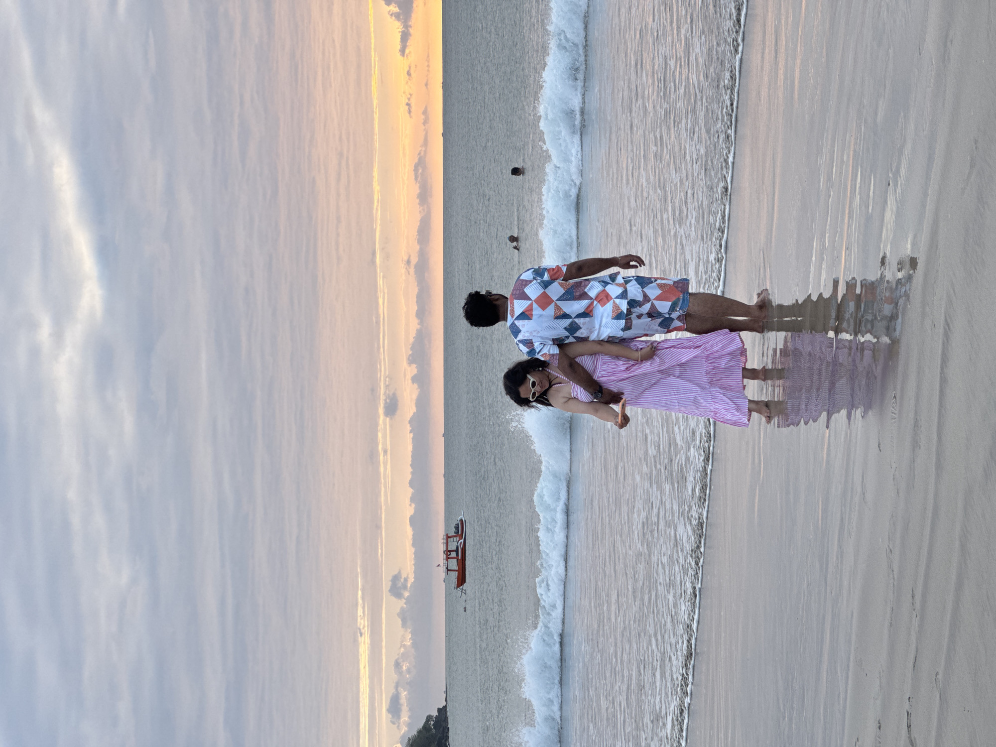This is a full-body, rear-view shot of a couple standing on a beach in Kuta Selatan, Indonesia, during sunset. The sky is filled with soft, layered clouds in shades of grey, white, and a hint of pink and orange from the setting sun. The ocean in the background has moderate waves breaking on the shore, and several small boats are visible in the distance.
The man is wearing a white t-shirt with a geometric pattern of red, blue, and white squares and triangles, and matching patterned shorts. He has dark hair and is standing with his right arm around the woman. The woman is wearing a pink and white striped maxi dress and white sunglasses. Her left arm is holding the man's arm, and her right hand is holding a smartphone, as if taking a photo or video. She has long, dark hair. Both are barefoot, with their feet in the shallow water at the edge of the sea. The wet sand reflects the couple and the colorful sky. In the middle ground, two people are visible swimming in the ocean.