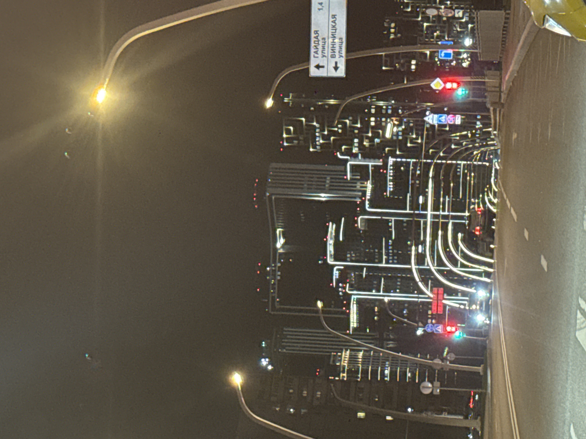 This vertically oriented image captures a bustling cityscape at night, likely in Moscow, Russia, as indicated by the visible text. The scene is dominated by a dark asphalt road on the right side, with faint white lane markings. Numerous streetlights, mounted on tall poles, extend along the road, casting bright yellow-orange light that creates noticeable lens flares.
In the midground and background, the city comes alive with countless illuminated buildings, their windows and architectural outlines forming intricate grid-like patterns and bright lines against the dark sky. Traffic lights are visible, with one showing both red and green signals, and another displaying a red light, indicating active traffic flow.
A prominent blue road sign with white Cyrillic text is visible on the left side of the road. It directs traffic, featuring an upward arrow pointing to "ГАЙДАЯ улица" (Gayday street) and a downward arrow for "ВИННИЦКАЯ улица" (Vinnitskaya street). The number "1.4" is also present on the sign, likely denoting a distance in kilometers. The night sky is dark and appears clear, with no visible precipitation or fog. The overall impression is one of an active, well-lit urban environment after dark.