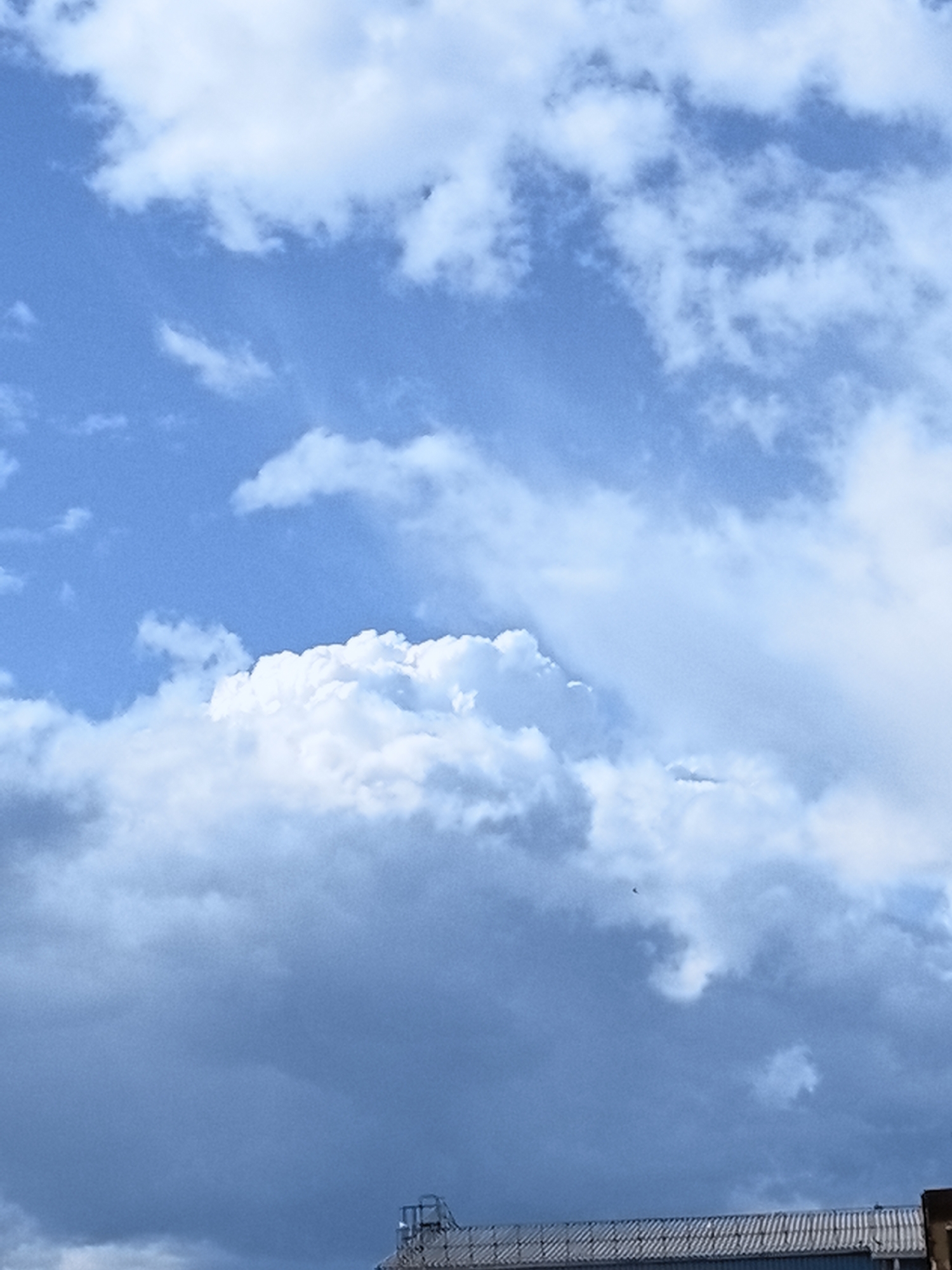 The image is an outdoor shot of a cloudy blue sky with a dark, ominous cloud formation taking up the lower portion. A sliver of a rooftop structure, possibly part of a building or industrial facility, is visible at the very bottom. There are no people or animals present in the frame. The scene suggests a potentially stormy or overcast weather condition. The location is identified as Ivanovo, Russia.
