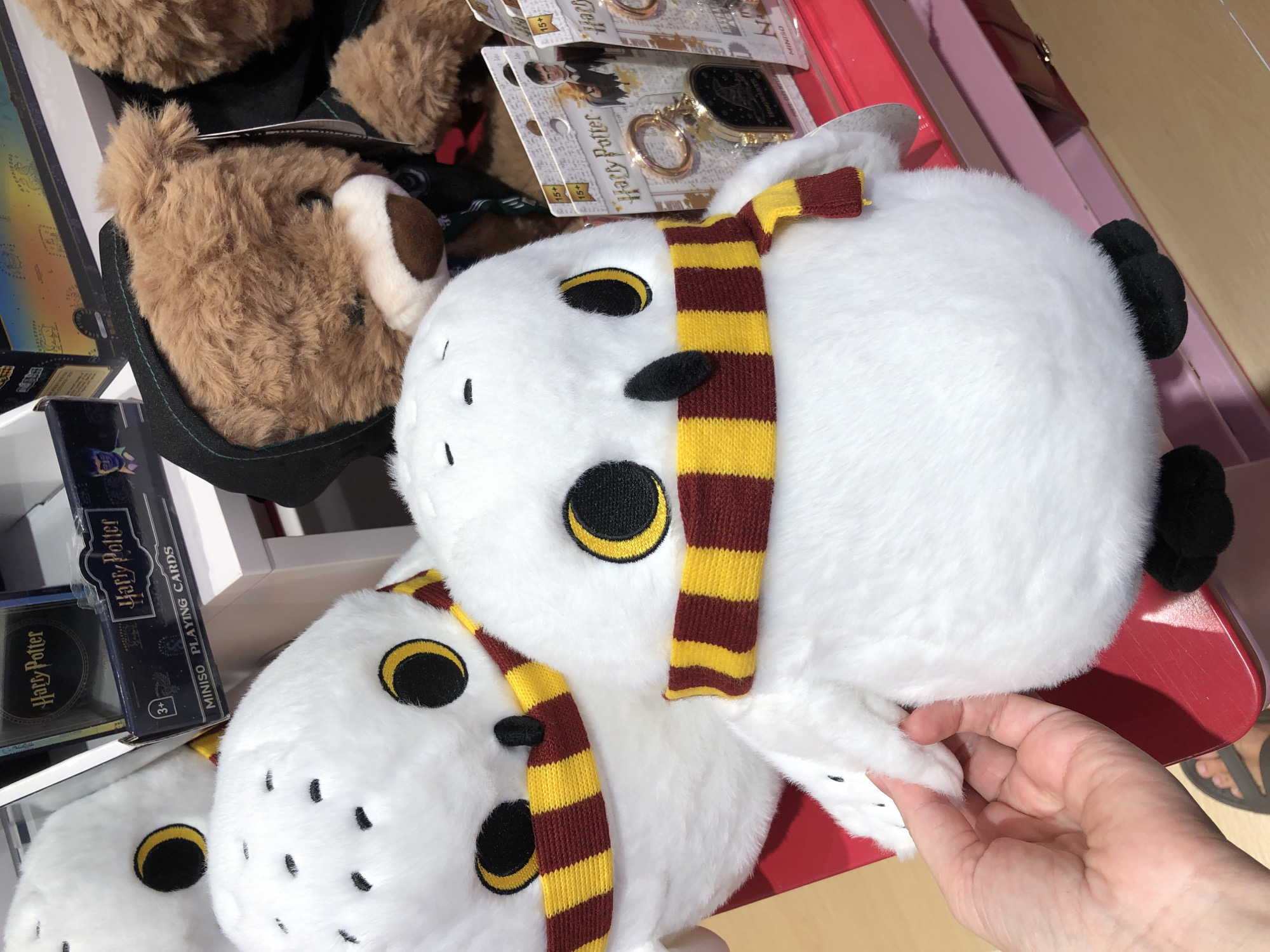 The media file is a close-up, top-down shot of several plush toys, likely in a store. The most prominent subjects are two white plush owls, one larger than the other, both wearing striped red and yellow scarves, characteristic of Harry Potter merchandise. The larger owl is positioned in the foreground, its body filling much of the frame. To its left, a portion of a brown plush bear or dog is visible, wearing a dark green collar.
In the background, several items of Harry Potter merchandise are displayed, including boxes of playing cards and keychains, indicated by the visible text "Harry Potter" and "MINISO". The products are arranged on white shelves and a red surface, suggesting a retail environment. A human hand is visible on the right side of the frame, gently touching the larger owl's wing, hinting at interaction with the products.
The lighting is bright, casting shadows and indicating an indoor setting, possibly during daylight hours. The overall mood is one of whimsical fandom and consumer interest, with the focus on the popular Harry Potter franchise. The location context provided is Đà Nẵng, Vietnam, which helps to contextualize the type of retail environment where such items might be found.