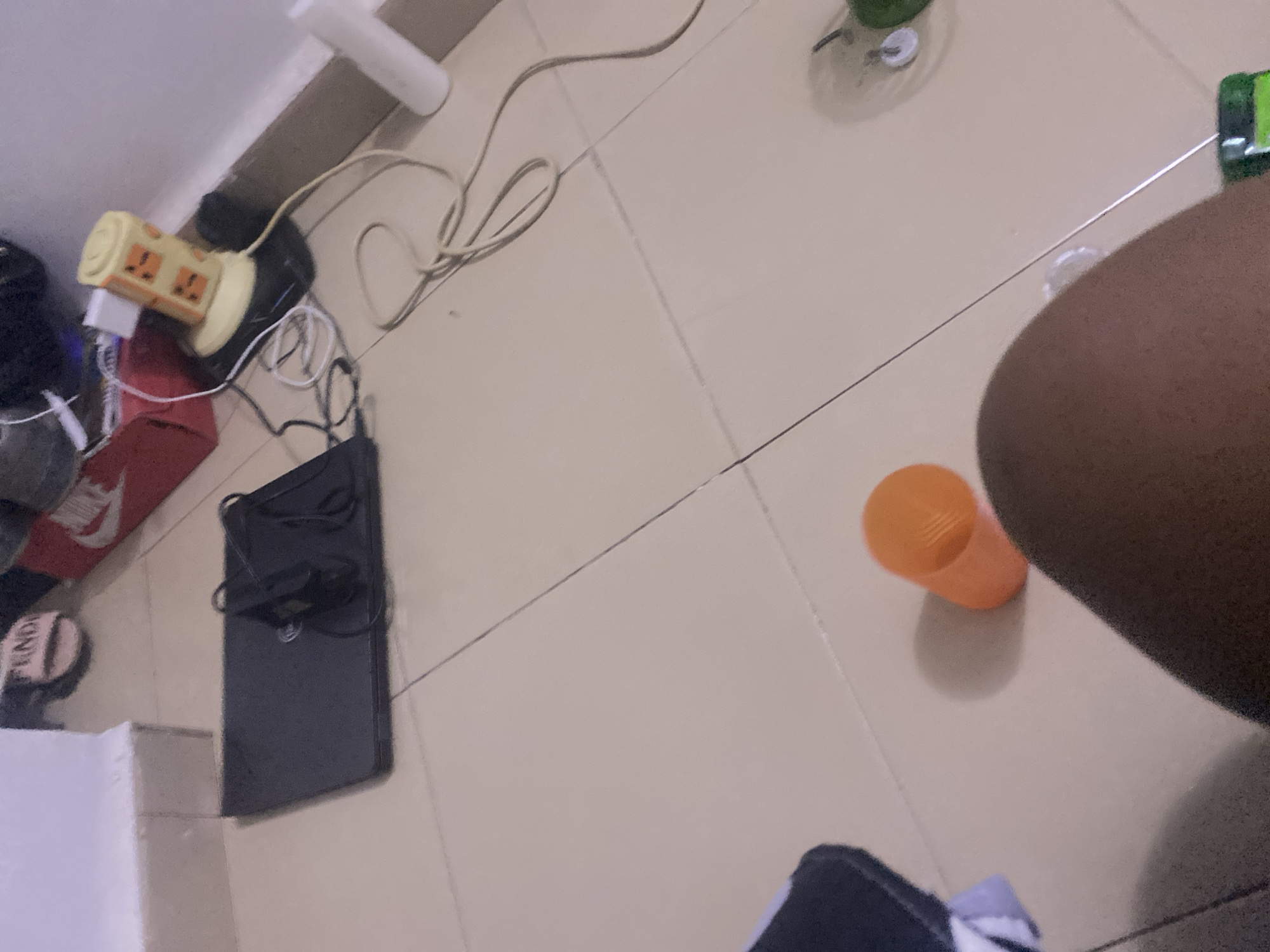 The image depicts a domestic interior scene, likely a bedroom, in Howayi, Nigeria. The floor is tiled in a light beige color, with visible grout lines. In the upper left, a white upholstered stool or low table is partially visible. A black laptop is open on the floor in the center of the image, with its charger plugged in and coiled nearby. To the right of the laptop, a yellow cylindrical object with orange square accents, resembling a power strip or adapter, is plugged into a black device. Various cables are strewn across the floor, connecting to the devices. In the foreground, a person's leg and knee are visible, along with a bright orange plastic cup lying on its side. In the background, out of focus, are what appear to be red and white shoeboxes, one with the Nike logo. A white spray bottle is visible near the right edge of the frame. The overall impression is one of a somewhat cluttered, lived-in space with electronic devices and personal items.