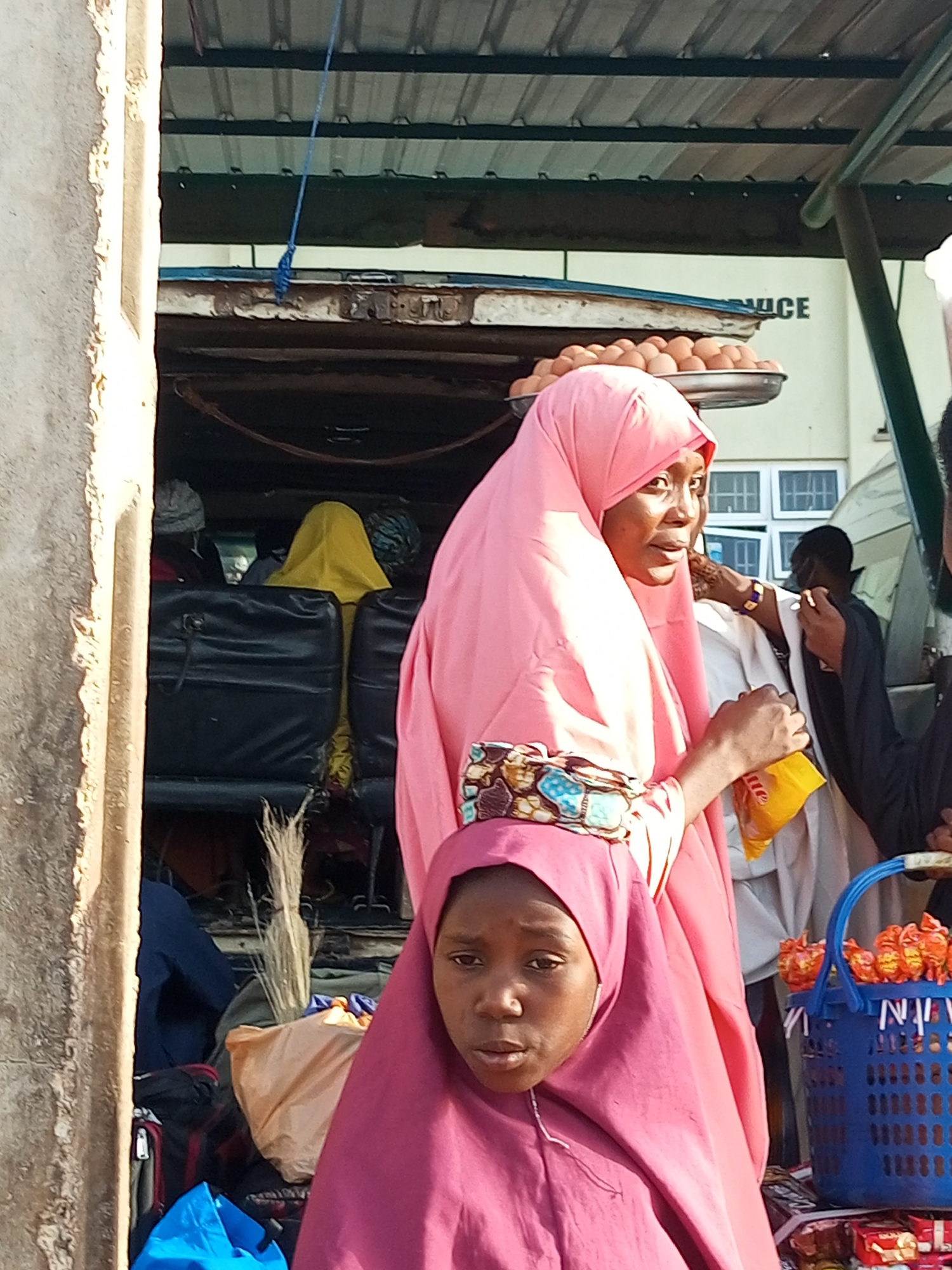 The image depicts a street scene in Gombe, Nigeria, likely at a market or transportation hub. In the foreground, two young females are visible, both wearing pink hijabs. The girl on the left is closer to the camera, with her face partially obscured by her hijab. The girl on the right is slightly behind and to the left of the first, her face more clearly visible as she looks towards the right of the frame.
Behind them, in the middle ground, several individuals are seen through an opening, possibly the interior of a vehicle or stall. One person is wearing a yellow hijab, another a grey head covering, and others are less distinct. Black seats of what appears to be a vehicle are visible.
In the background, a tray filled with eggs is balanced on top of something, suggesting a vendor. Further behind, a building with windows is visible, and the word "ICE" is discernible on its facade, possibly indicating a business name. A blue basket filled with orange-wrapped items, likely sweets or snacks, is placed on a surface in the right foreground, next to some other packaged goods. A bundle of straw or reeds is leaning against some luggage in the middle ground. The overall environment suggests a busy, outdoor commercial area.