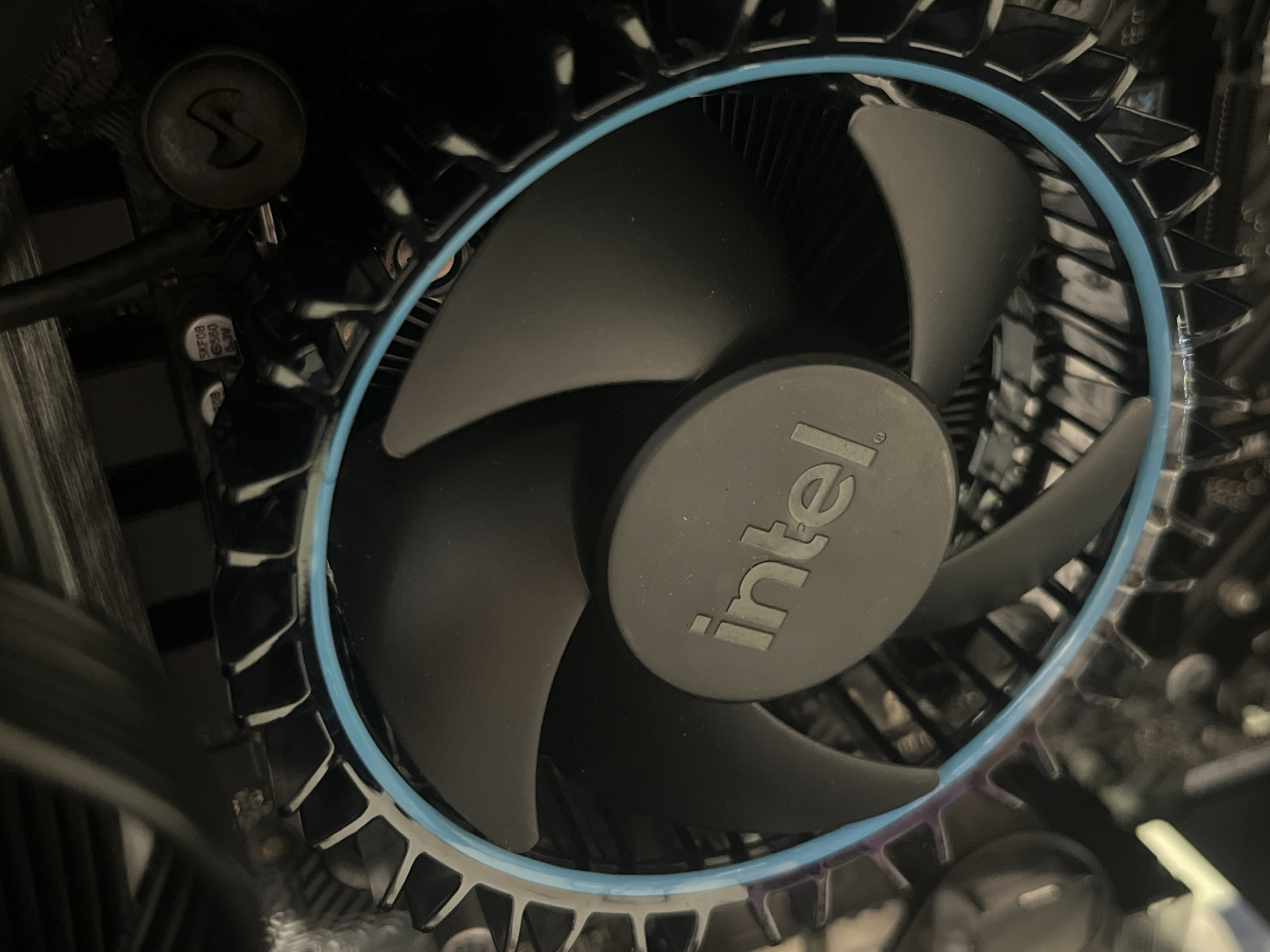 The image shows a close-up of an Intel CPU cooler installed inside a computer case. The cooler features a black fan with the Intel logo on the center hub, surrounded by a heat sink with a blue accent ring. The environment is inside a computer case, with glimpses of other components visible around the cooler. The location is Unknown, Unknown.
