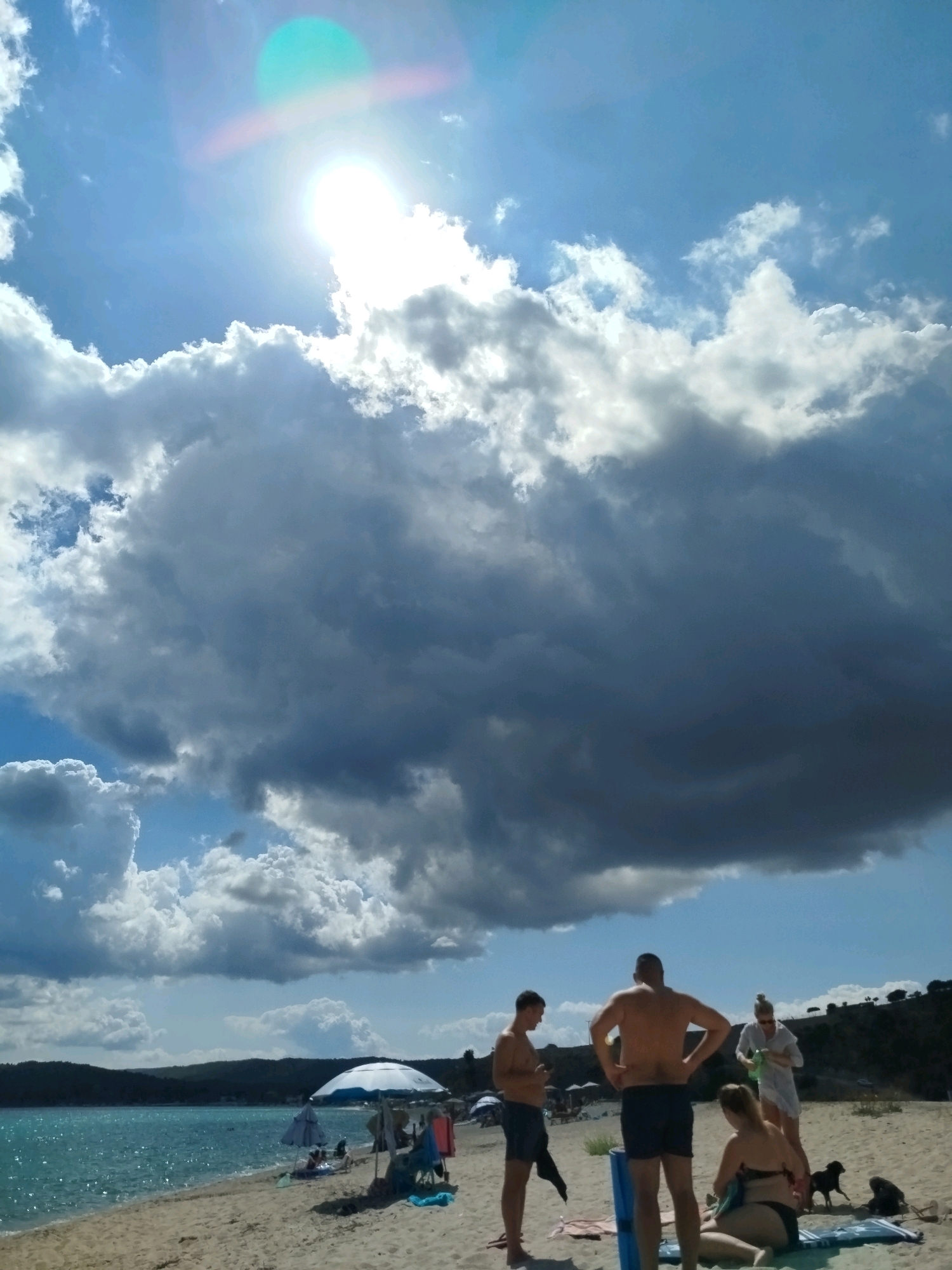 The image depicts a beach scene in Sithonia Municipal Unit, Greece. The sky is partly cloudy with a bright sun, casting a lens flare on the upper left portion of the image. In the foreground, four individuals are on the sandy beach. Two men and one woman are standing, and another woman is seated. One of the standing men is wearing blue swim trunks, the other is wearing black swim trunks. The woman in the white cover-up appears to be holding something green. A black dog is visible near the seated woman. The beach is situated alongside a turquoise body of water. In the middle ground, beach umbrellas and chairs are set up. Beyond the beach, a tree-covered hillside forms the background, with a clear blue sky overhead.