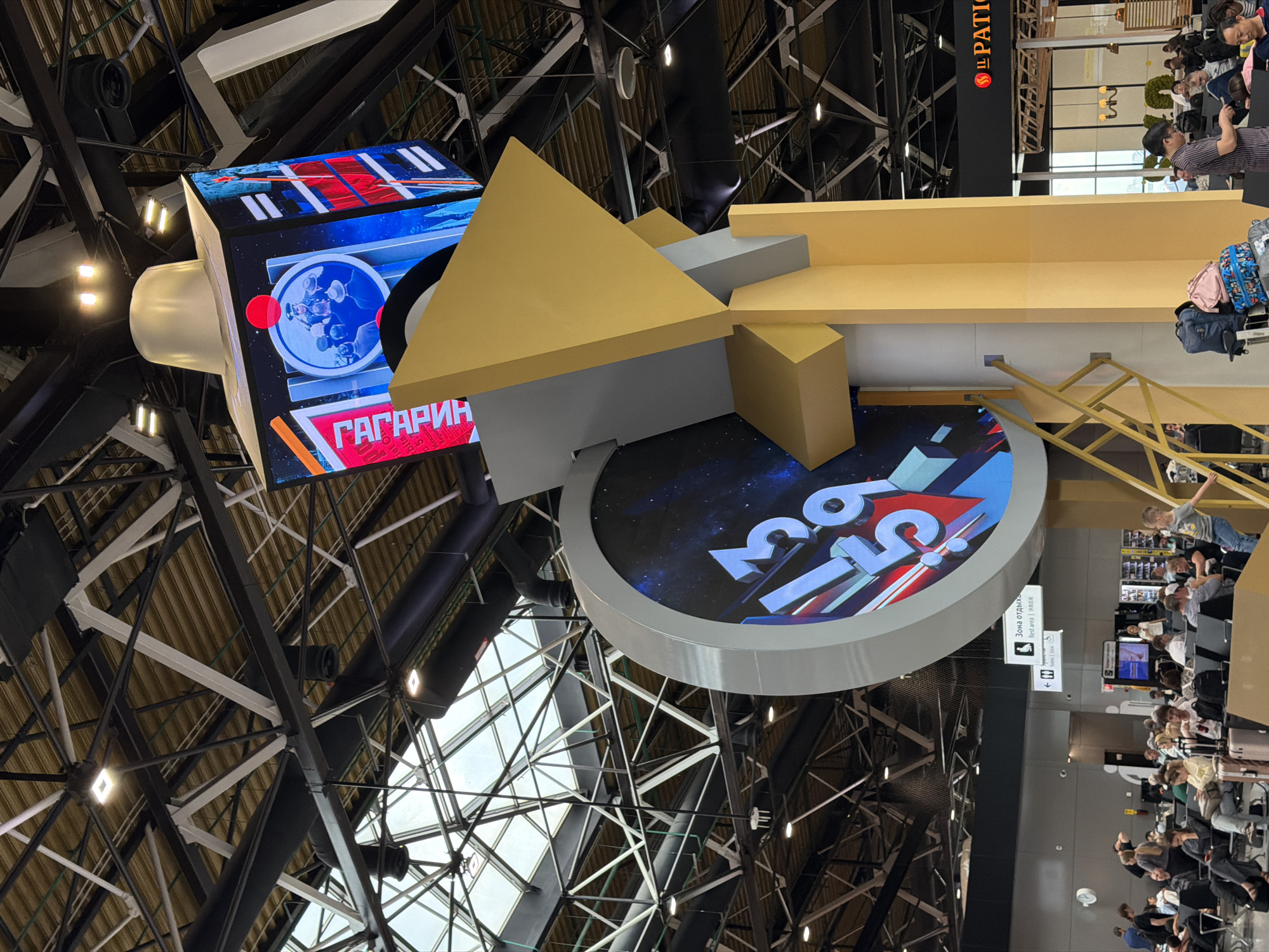 This image depicts the interior of a modern building, likely an airport or large public space, in Khimki, Russia. A prominent feature is a large, abstract sculpture with a digital clock displaying "13:59" on a circular face. Above this, a multi-faceted digital screen shows various images, including text that reads "ГАГАРИН" (Gagarin) and a circular image with a man in a military uniform. The ceiling is a complex network of black and white metal beams and ductwork, with skylights allowing natural light to enter. In the foreground, a group of people are seated in a waiting area, with luggage visible. There are signs indicating "Зона отдыха" (Rest Area). The overall environment appears to be well-lit and spacious.