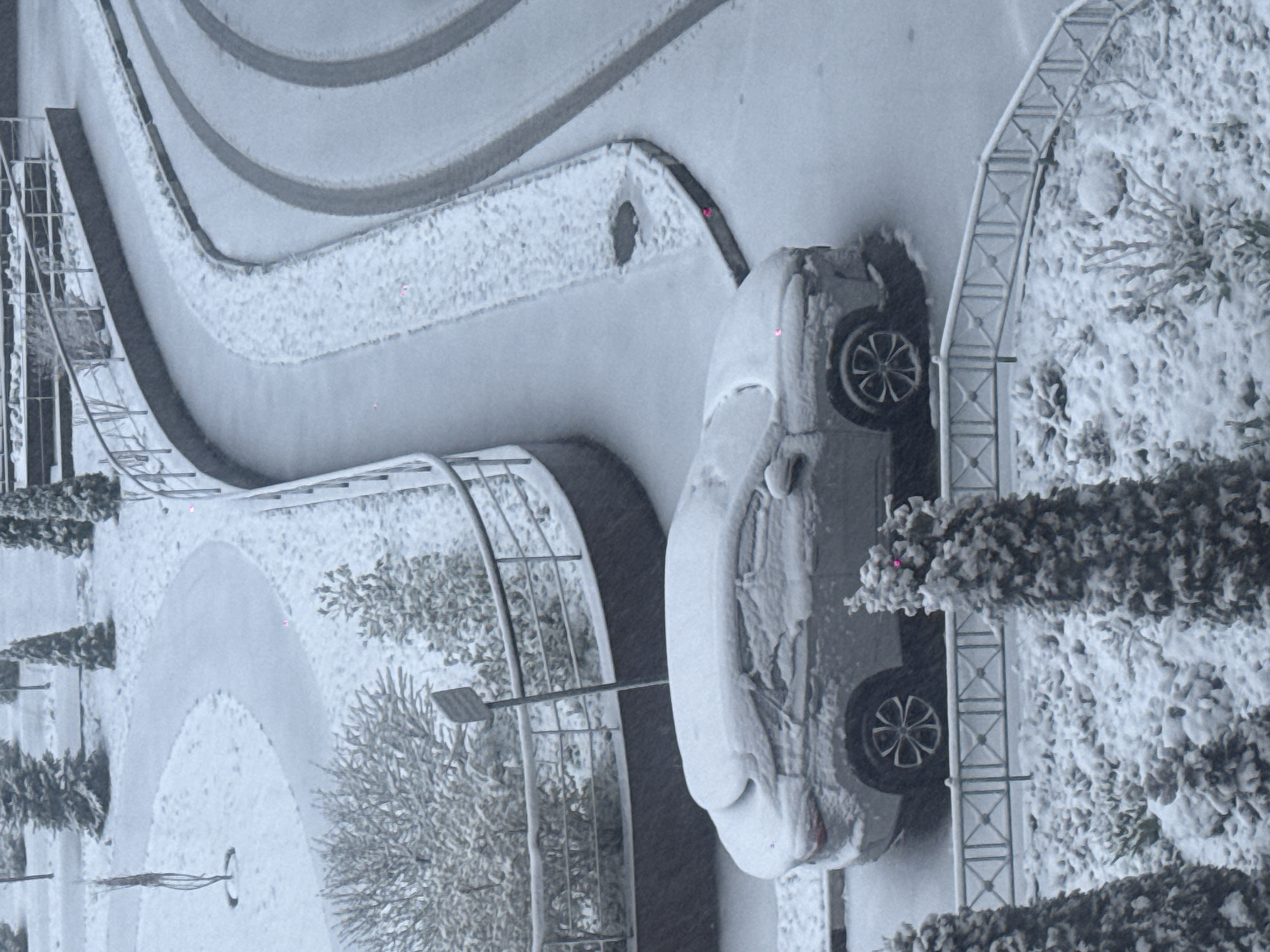 The image depicts a snow-covered scene in Khimki, Russia. A car is parked on a street, covered in snow. Adjacent to the street, there is a walkway with railings, also covered in snow. Bushes and trees are visible, heavily laden with snow. The environment appears to be a residential area.