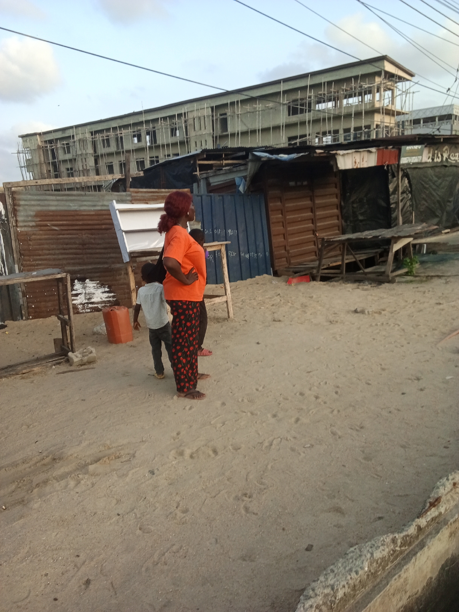 The scene is set outdoors in Ado, Nigeria, characterized by a sandy terrain and makeshift structures. A woman with reddish-brown hair, wearing an orange t-shirt and patterned pants, stands with her hand on her hip. A young boy is visible next to her. A larger building under construction can be seen in the background. Various temporary shacks constructed of materials like corrugated metal and dark tarpaulin line the foreground. Electrical wires stretch across the sky.