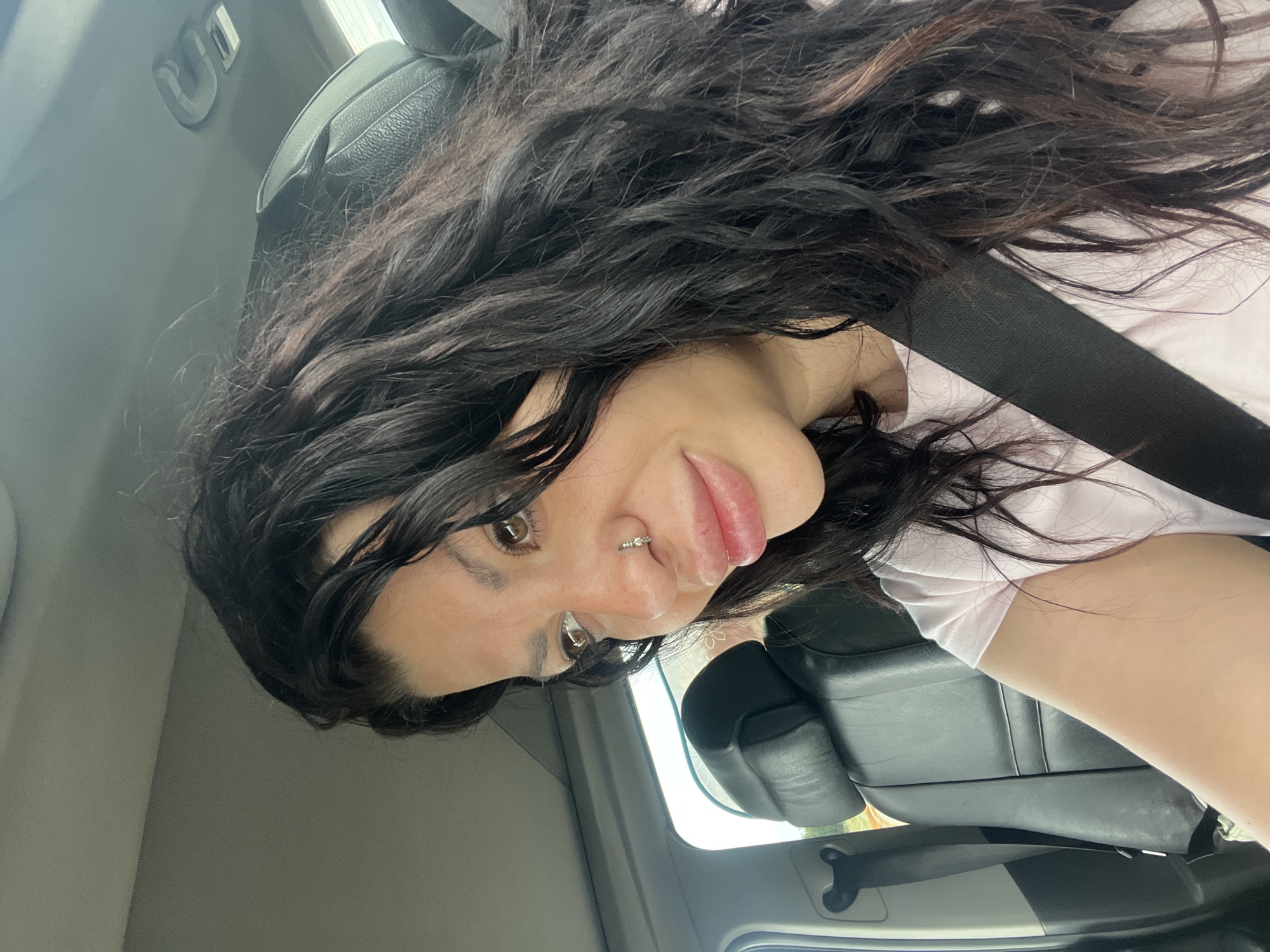 The image is a selfie of a person with dark, wavy hair and a light complexion. They are wearing a pink shirt and a seatbelt, suggesting they are in a vehicle. The background shows the interior of a car, with black leather seats and a grey interior trim. The location of Stryama, Bulgaria cannot be confirmed based on the image alone.