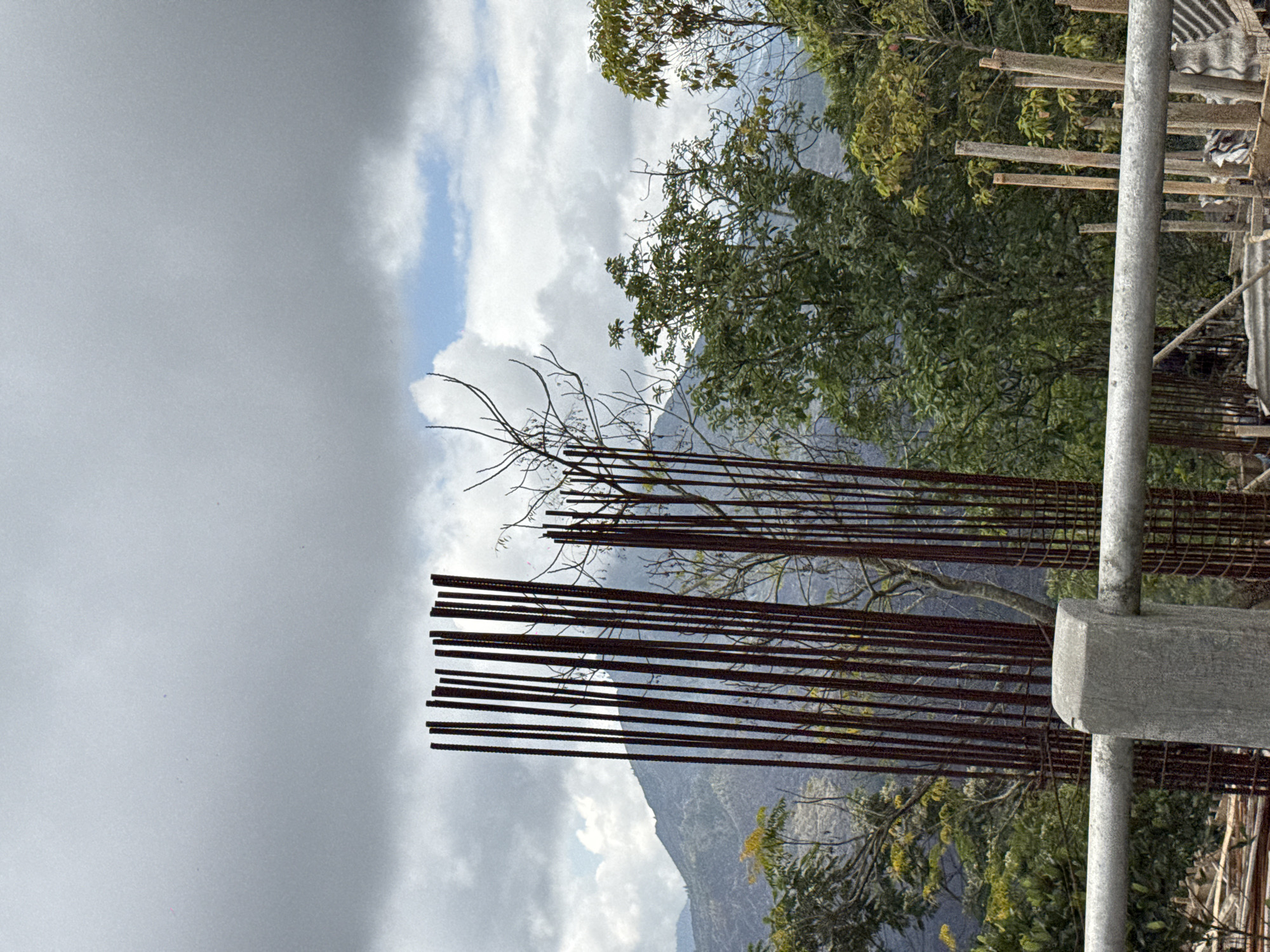In Kintamani, Indonesia, a construction site is partially visible against a backdrop of lush green hills and a cloudy sky. In the foreground, a concrete pillar supports a silver railing, suggesting a vantage point. Behind the railing, a cluster of rust-colored steel rebar stands vertically, indicating ongoing construction. Beyond the rebar, dense green foliage of trees dominates the midground, with some bare branches interspersed. In the background, a majestic mountain rises, partially obscured by clouds and mist. The sky is a mix of gray clouds and patches of blue, with hints of sunlight breaking through. The overall scene captures the raw state of construction against the serene natural beauty of Kintamani.