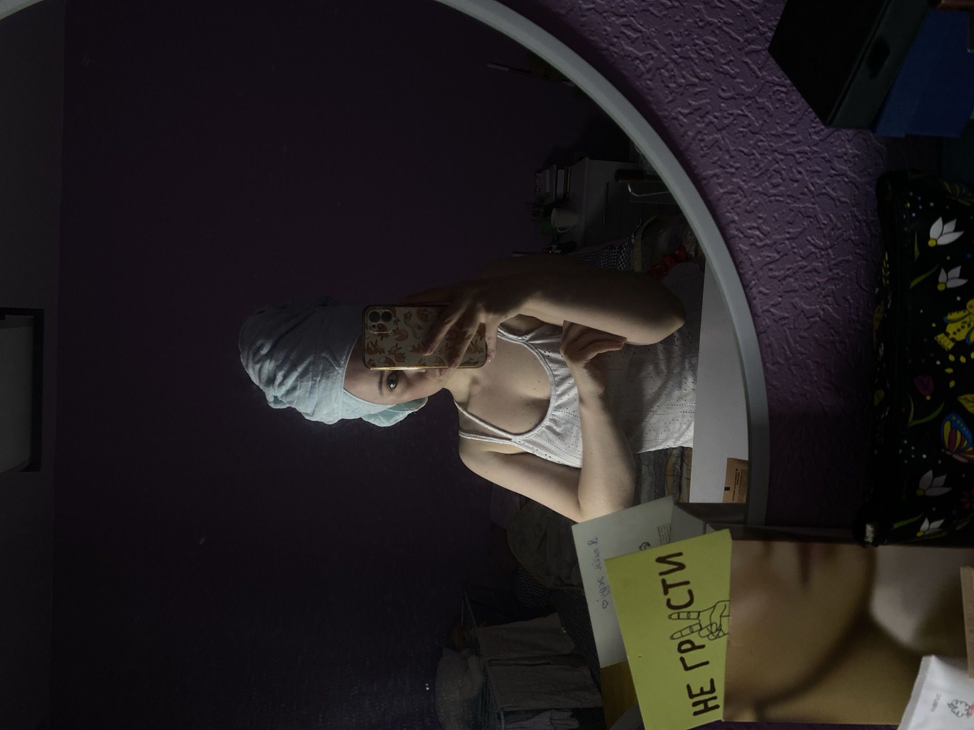 An indoor mirror selfie captures a young woman, likely in Saint Petersburg, Russia, after a shower or bath, as indicated by a light-colored towel wrapped around her head. She is wearing a white or light-colored tank top and holds a smartphone with a distinctive fox-patterned case to take the photo. Her gaze is directed at the camera through the mirror, with a neutral expression.
The mirror, framed in light grey, reflects a dimly lit room with dark purple, textured wallpaper. Various personal items are visible in the reflection, including what appears to be a white cabinet or shelves with clutter behind the woman. To the right, outside the mirror's reflection, is a dark fabric with colorful floral and butterfly patterns, possibly part of a bedspread or pillow.
In the foreground, partially obscuring the bottom right of the mirror, is a yellow rectangular card with handwritten Russian text: "НЕ ГРУСТИ," which translates to "DON'T BE SAD." A simple drawing of a hand making a gesture (possibly "rock on" or "peace") is next to the text. Another white piece of paper next to the yellow card has faint handwriting, possibly containing a heart symbol and more Russian text. The overall scene suggests a private, personal moment in an everyday setting, possibly in the evening given the subdued lighting.
