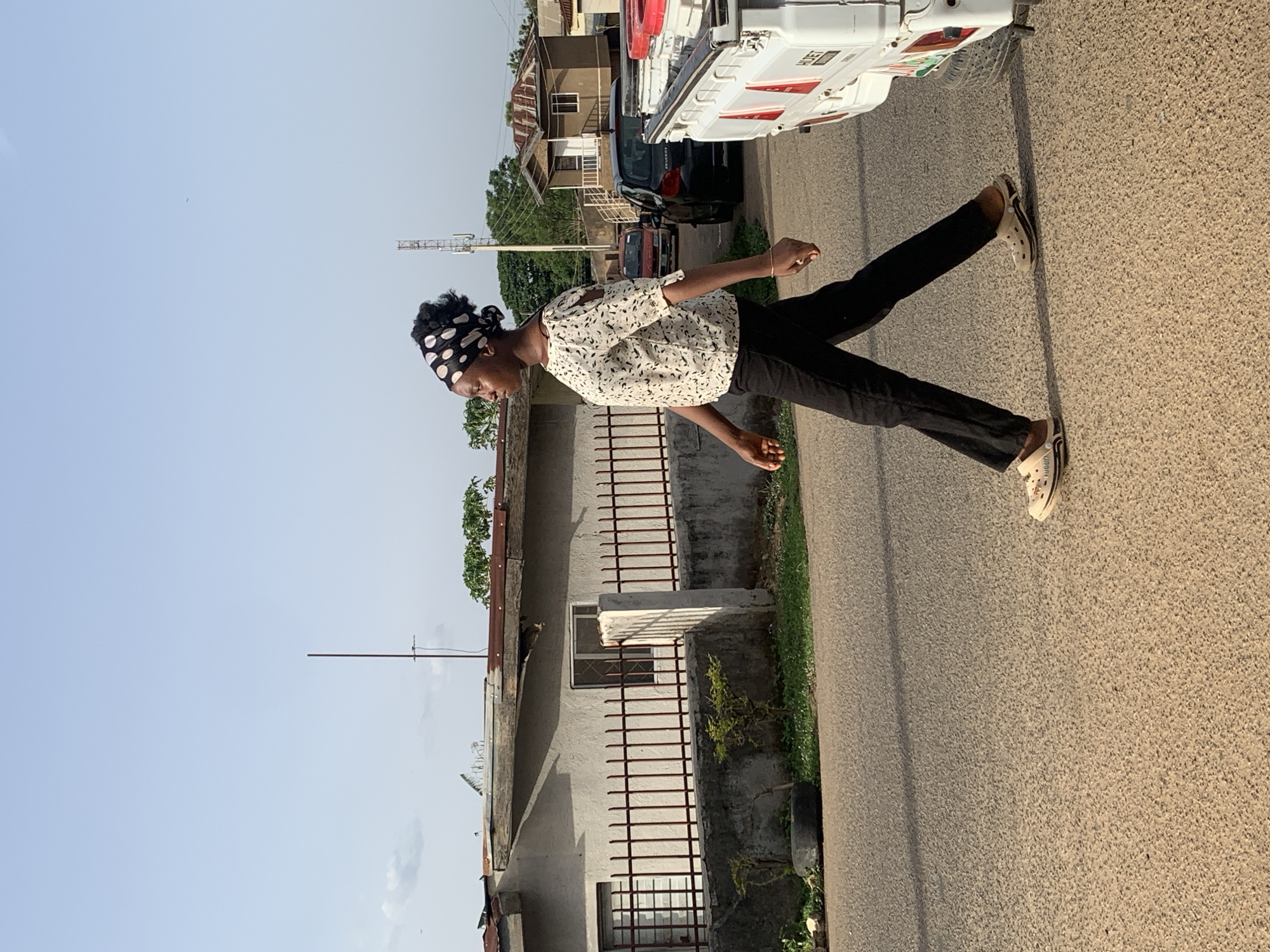 A young woman stands on the side of a road in what appears to be Jos, Nigeria. She wears a white patterned blouse, black pants, and cream-colored slip-on shoes. A black and white polka-dot headscarf covers her hair. She is positioned on the right side of the image, next to a paved road. Behind her is a building with bars on the windows and a rusty roof. A utility pole stands near the building. A white truck is partially visible in the background, along with a few other vehicles and some houses. The sky is visible in the upper left of the image.