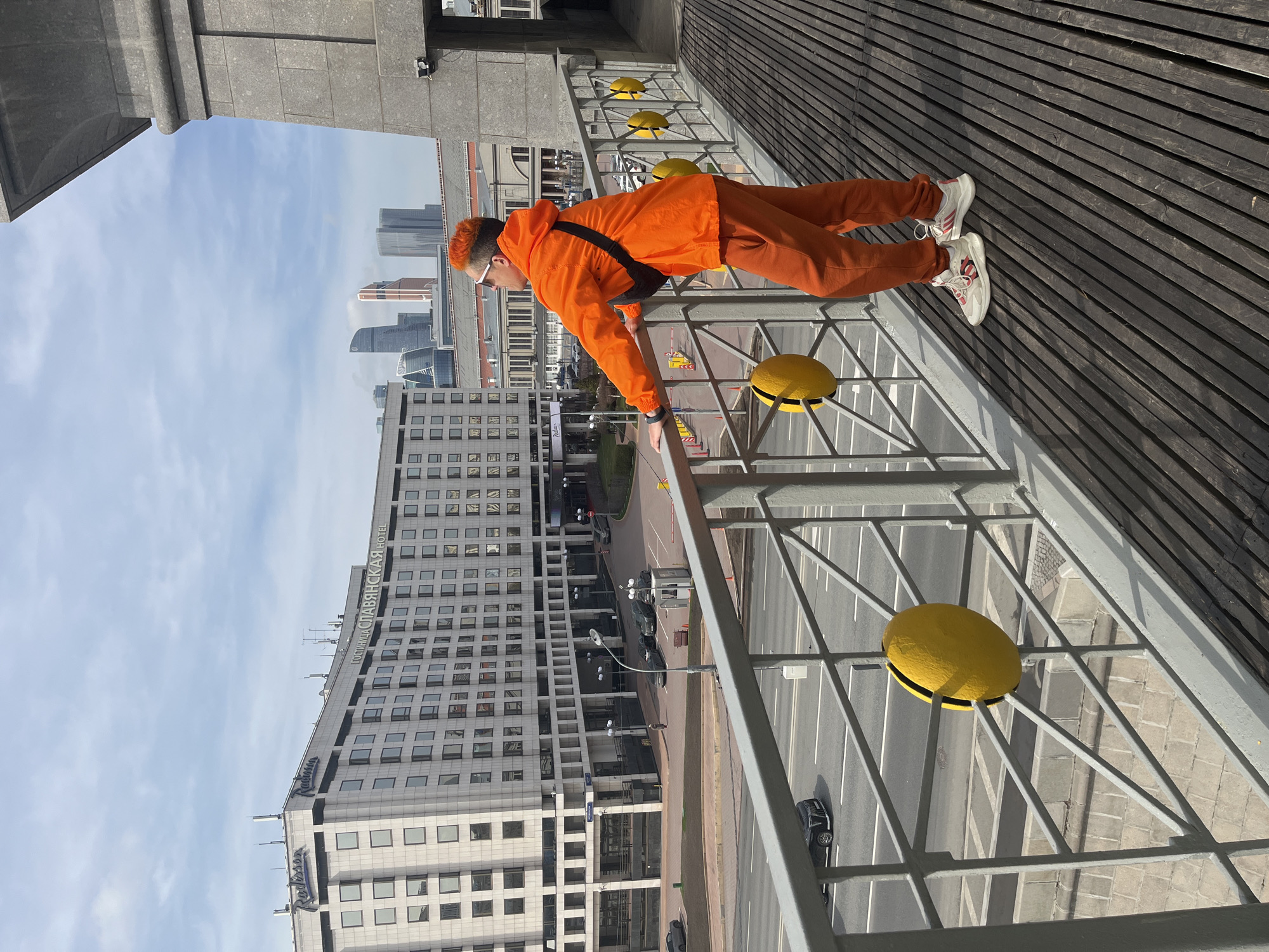 A person in an orange jacket and pants leans over a bridge railing in Moscow, Russia. The Radisson Slavyanskaya Hotel is visible below, with Moscow City skyscrapers in the background. Yellow discs are attached to the bridge's railing. The person wears white sneakers and a black crossbody bag. The bridge has a wooden walkway. A street with moving cars is below. The sky is partly cloudy.