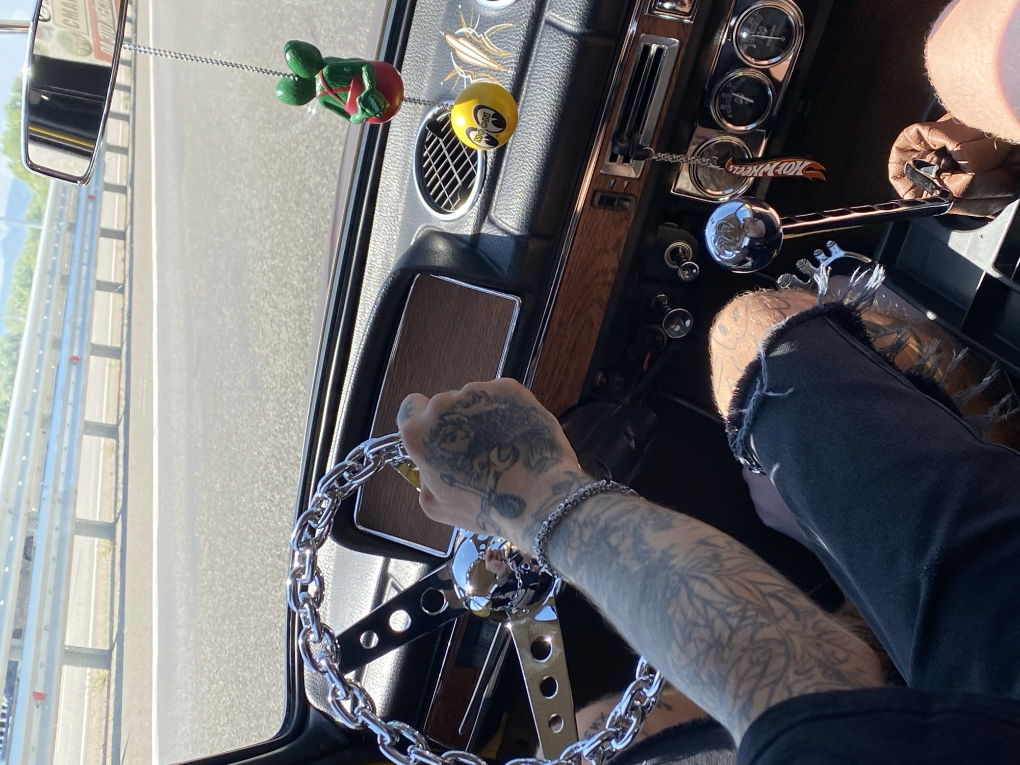 A first-person perspective captures a person driving a car on a sunny day, likely on a highway. The car's interior features a chrome steering wheel with a large chain design and a wooden trim on the dashboard. A tattoo is visible on the driver's arm and hand, which is gripping the steering wheel. Hanging from the rearview mirror is a green bobblehead figure and a yellow ball-shaped air freshener with a "MOON" logo. Additional car accessories include a Hot Wheels keychain hanging from the gear shift. The exterior view through the windshield shows a highway with a metal guardrail and trees in the distance. The image is likely taken in the city of Goryachiy Klyuch, Russia, based on the license plate visible in the rearview mirror, which appears to be a Russian plate.