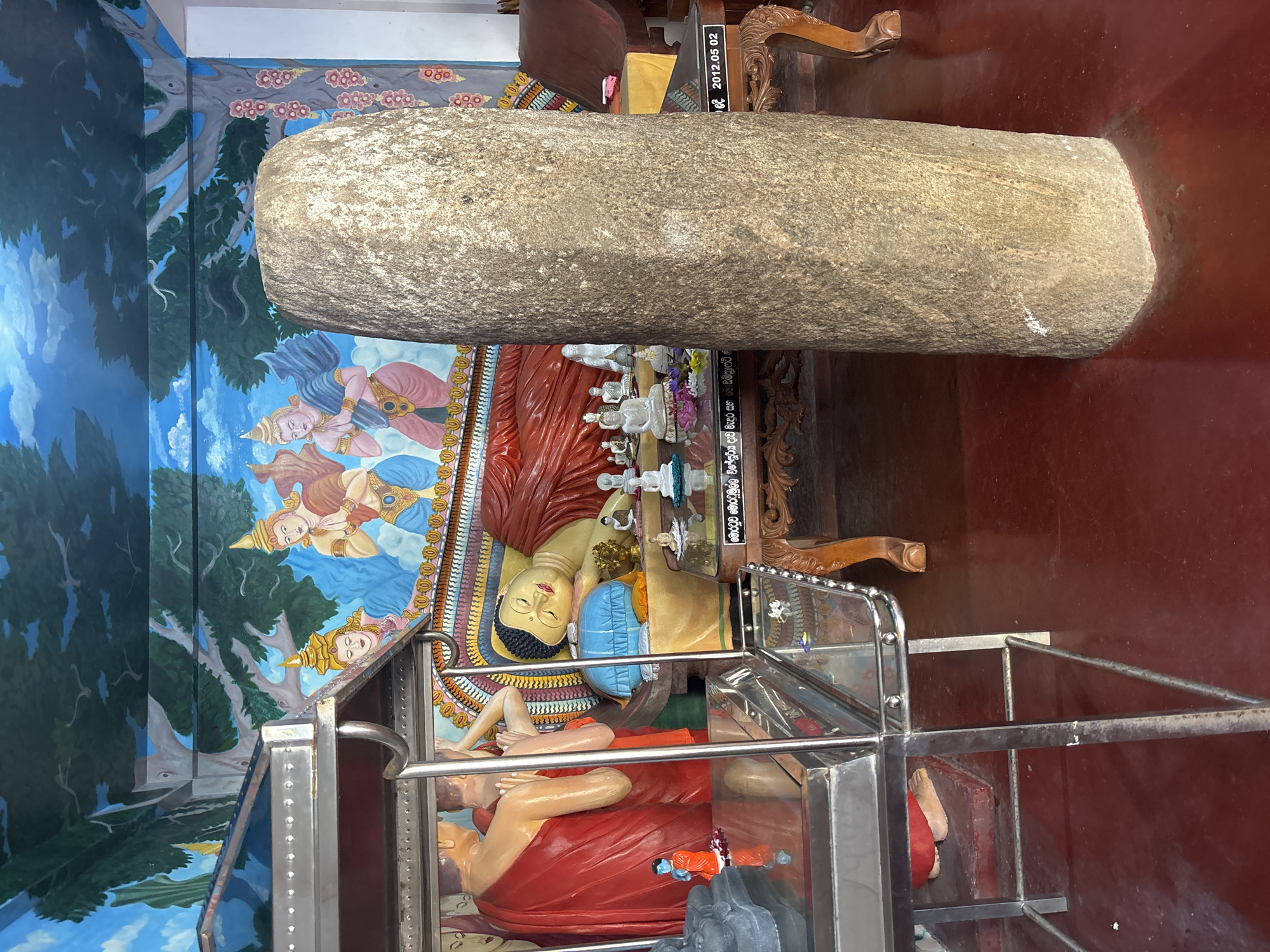 The image depicts a religious site, specifically a temple, possibly located in or near Dondra, Sri Lanka. A large, cylindrical stone object is the focal point, positioned over a glass-encased display of reclining Buddha statues. The statue is red and yellow. The scene is further adorned with a painted mural on the wall featuring figures with blue and yellow coloring, along with stylized trees. The flooring is a shade of red. A wooden chair is also present in the scene. A plaque reads "2012.05.02".