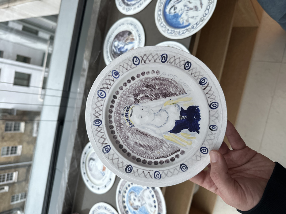 A white plate with a blue and brown drawing held in a hand. The drawing depicts a face with brown hair and blue details, surrounded by similar colors. Decorative circles surround the edge of the plate, along with other decorative patterns. Several other similar plates are behind it and to the left in the background, some with similar color schemes of blue and white. A window with a building beyond is to the left. A wooden surface is to the right of the plate.
