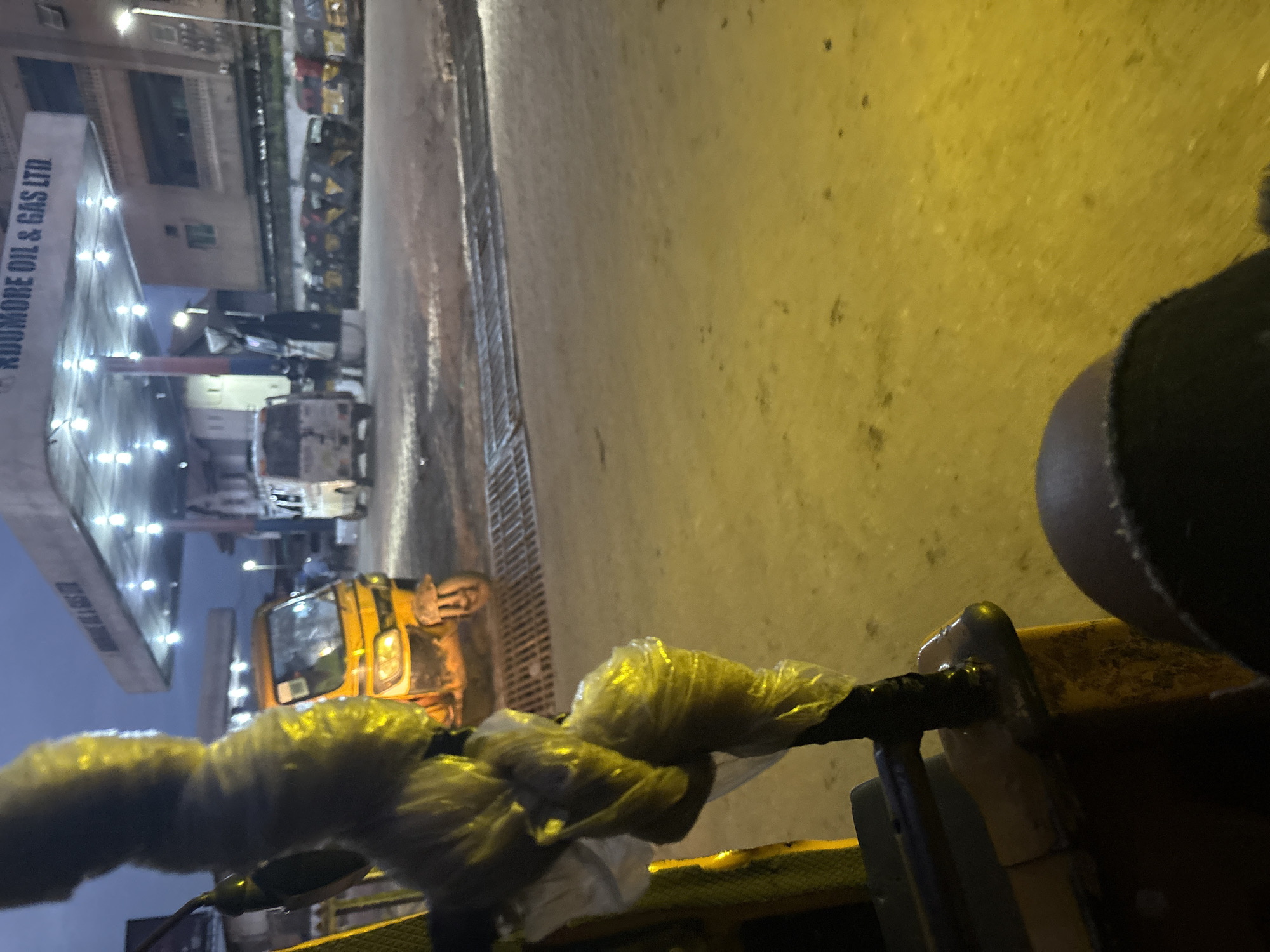 The image captures a scene at the Udoremore Oil & Gas Ltd. station in Onitsha, Nigeria, during what appears to be dusk or early evening. The foreground is dominated by the interior of a vehicle, likely a keke napep or auto-rickshaw, with its handlebars wrapped in translucent plastic. A yellow auto-rickshaw is parked to the left, its front wheel and part of its body visible. In the middle ground, a white van is parked near the gas station's canopy. Several other vehicles, including what appear to be more auto-rickshaws and possibly minibuses, are lined up further back. The gas station canopy, illuminated by overhead lights, is a prominent feature, with the "Udoremore Oil & Gas Ltd." signage visible. The ground is paved and wet, reflecting the lights. The sky is a dusky blue, suggesting a transition from day to night.