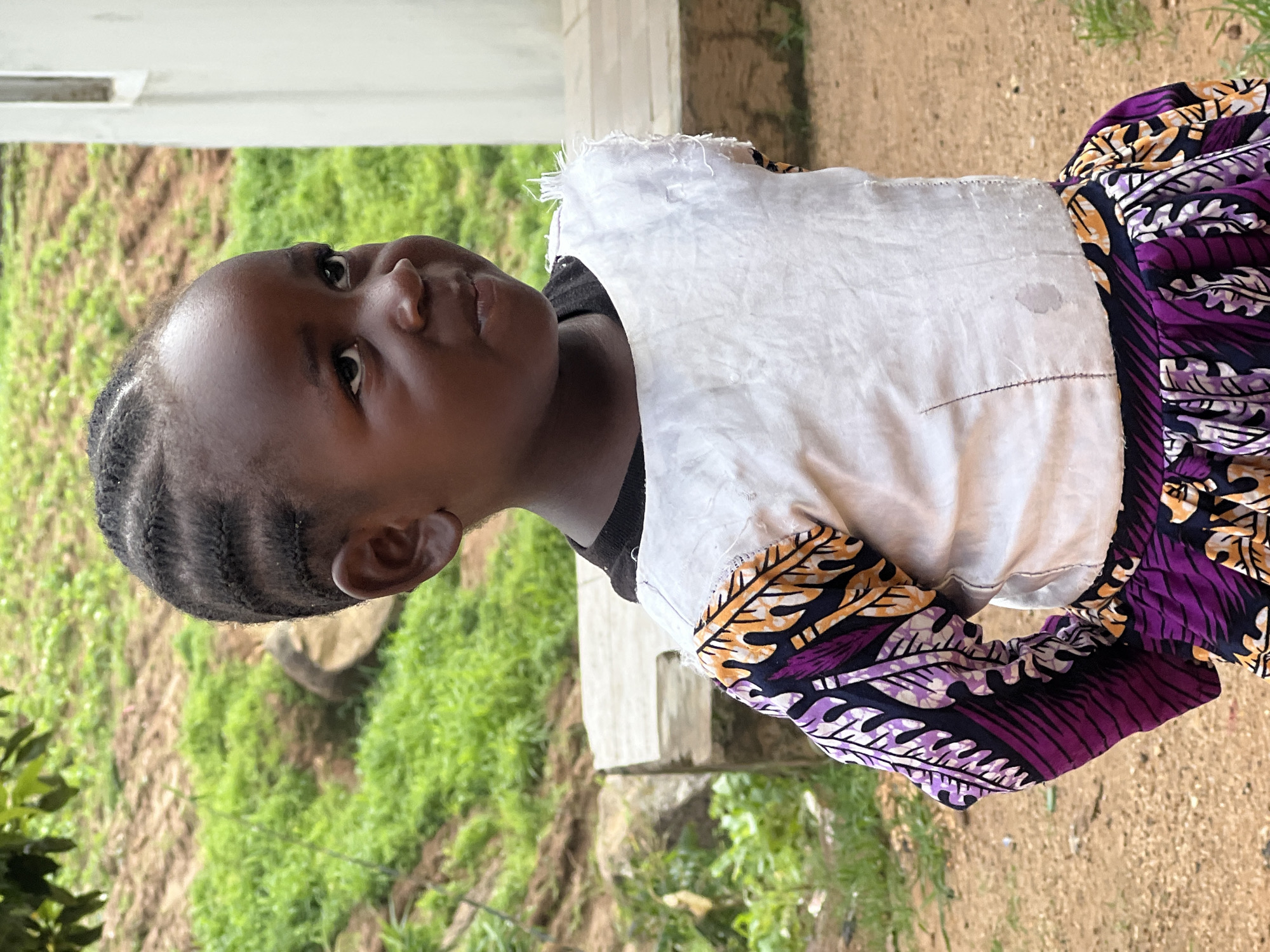 A young girl of African descent, with dark skin and hair styled in braids, is pictured from the chest up, looking upwards and to her left. She is wearing a black t-shirt layered with a white, roughly stitched vest, possibly homemade, with frayed edges at the top. The vest is overlaid with a colorful garment featuring a vibrant purple, black, and yellow patterned fabric, likely an Ankara print, with a leaf motif. The girl's expression is neutral, with her eyes open and looking slightly to the side. Her left ear is visible.
The setting appears to be outdoors, with a blurred background of lush green foliage and dry, earthy ground. A light-colored, possibly concrete, structure is visible behind her. The lighting suggests it is daytime, with soft illumination. The overall impression is that of an informal, possibly rural or community setting in Jos, Nigeria. There is no visible text in the image. The image is oriented vertically, with the subject facing the right side of the frame.
