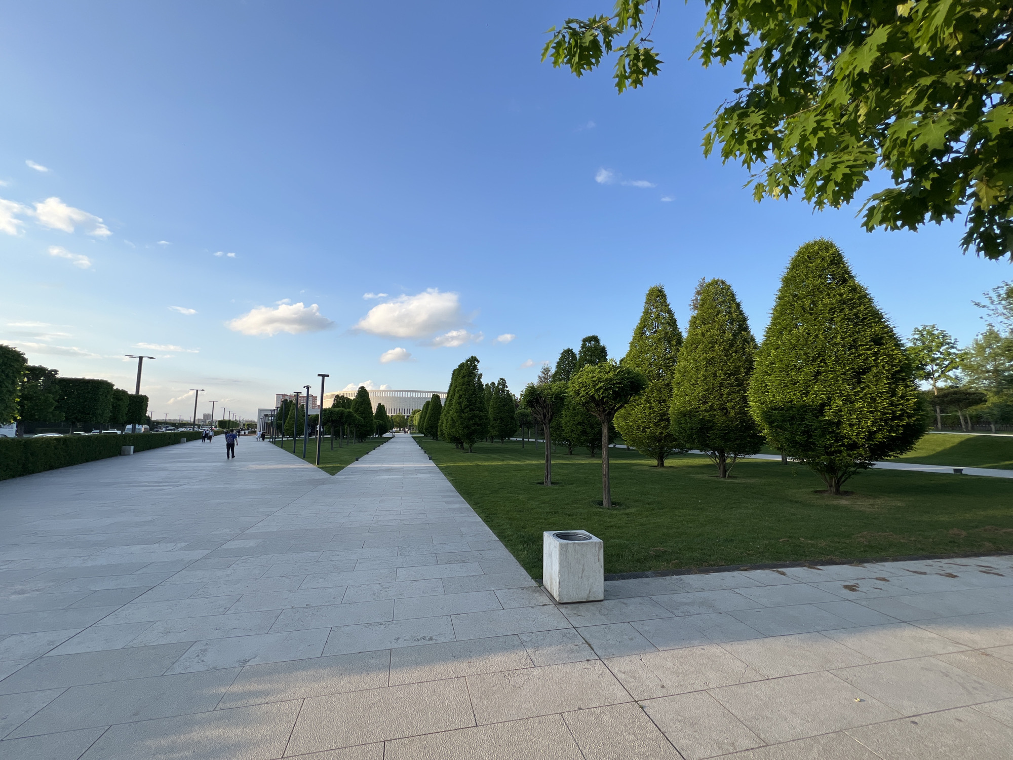 The image depicts a pathway in a park located in Krasnodar, Russia. The pathway is constructed of light-colored stone tiles. On one side, there's a green lawn with evenly spaced, cone-shaped evergreen trees and smaller trimmed trees. On the other side, a hedge runs along the pathway, beyond which there are buildings and parked vehicles. In the distance, a stadium is visible. There is a person walking along the left pathway in the distance. A concrete trash receptacle is beside the path on the right. The sky is blue with scattered clouds. The foliage on the upper right indicates the presence of another tree nearby, with its leaves partially obscuring the sky.