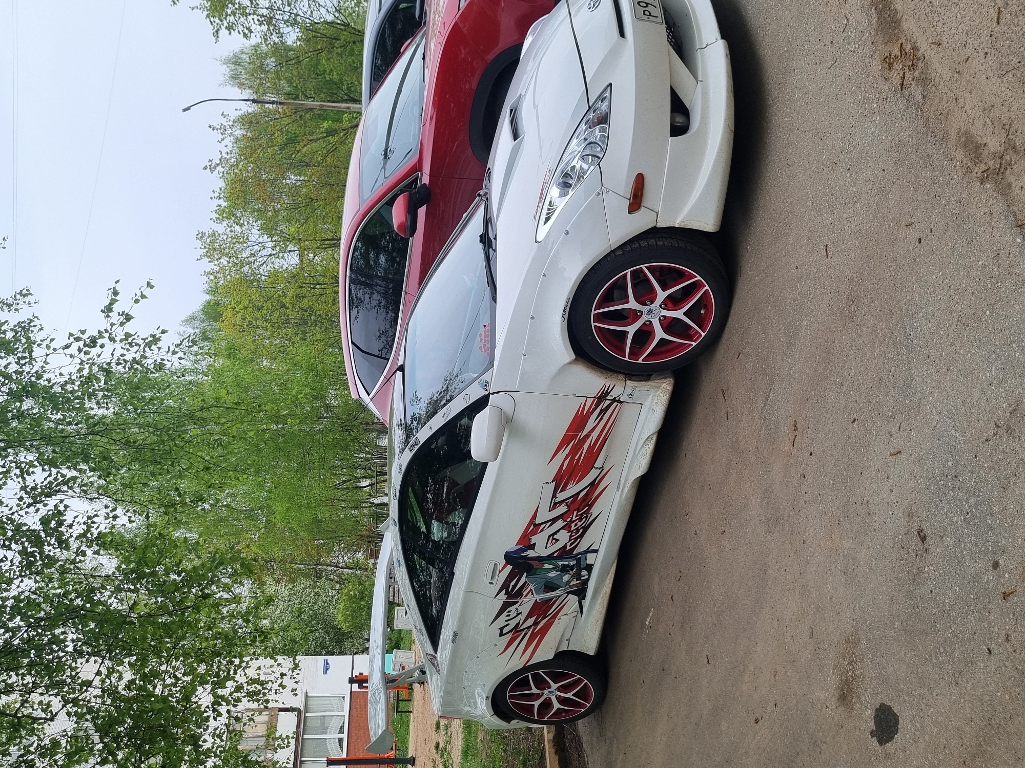 The image depicts a white Toyota Celica parked on a concrete surface. The car has custom red and white rims, a large spoiler, and anime-style graphics on the side. The image also shows a red SUV partially visible behind the Celica. In the background, there are trees and a building that reads "Ростелеком" in Russian. The location appears to be Chaikovsky, Russia. There are no people visible in the image. The image presents a static scene with parked vehicles.