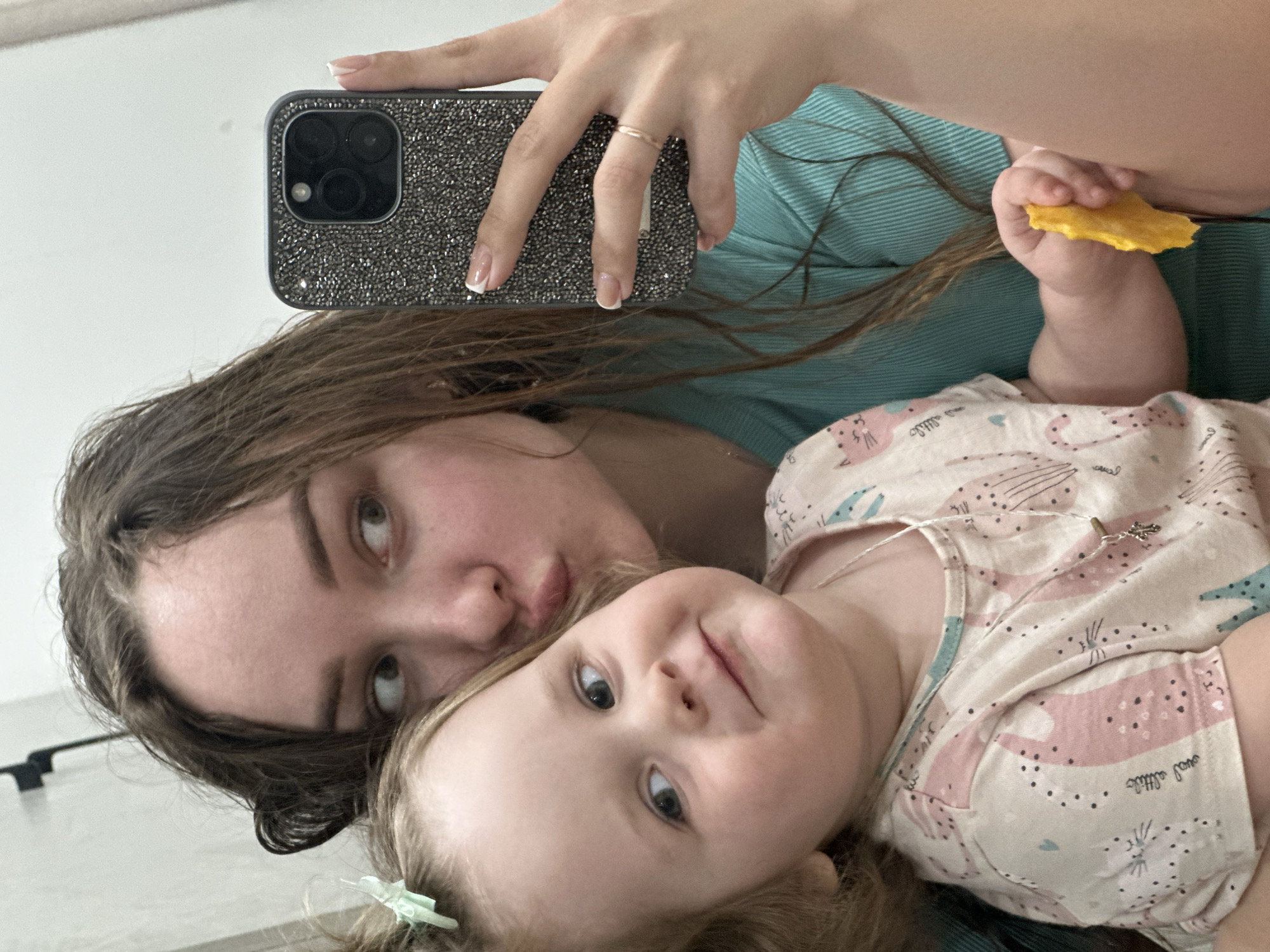 The image shows a selfie of a young woman and a toddler, likely a mother and child. The woman has long brown hair and is wearing a blue shirt. She is holding a smartphone with a sparkly case. The toddler is wearing a pink and white patterned outfit and a small hair clip. The child is holding a yellow object, potentially food. The background appears to be a plain wall, possibly indoors. There is no evidence suggesting the photo was taken in Chaikovsky, Russia.