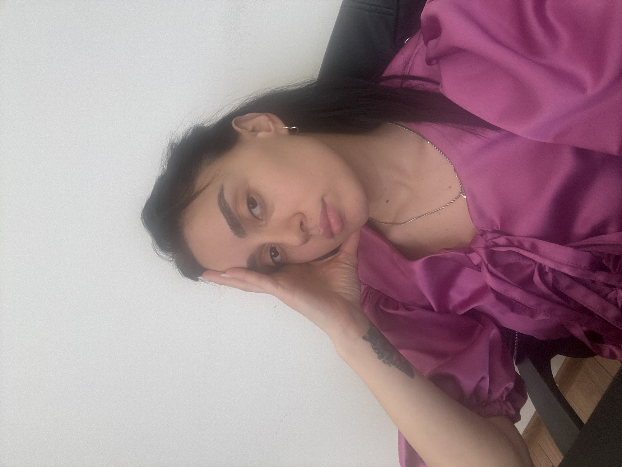 A medium, slightly rotated, eye-level shot depicts a woman indoors against a pale wall. She is seated, wearing a vibrant pink, silk-like garment with puffed sleeves. Her dark hair is pulled back, revealing a silver chain necklace and small hoop earrings. Her left hand is resting on her cheek, supporting her head. A visible tattoo adorns her left forearm. The chair she is seated in is black. The precise location in Tbilisi, Georgia, cannot be determined from this image.