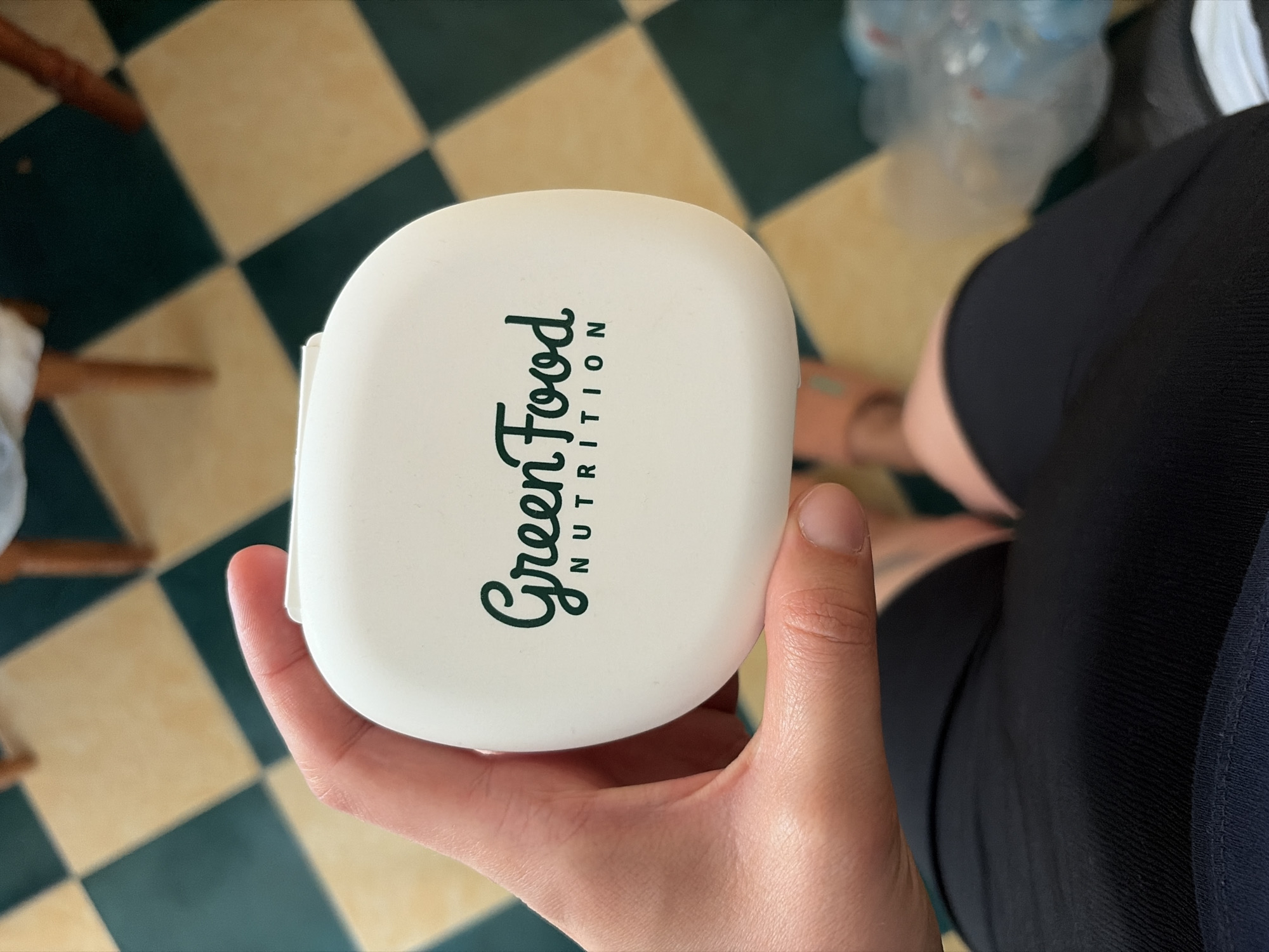 A person's hand, with visible fingernails, holds a white, rounded container with the logo "Green Food NUTRITION" printed in dark green text. The container has a slightly glossy finish. The background is a tiled floor with alternating yellow and dark green squares. In the background, out of focus, are the legs of a wooden chair and a plastic bag. The scene appears to be indoors.