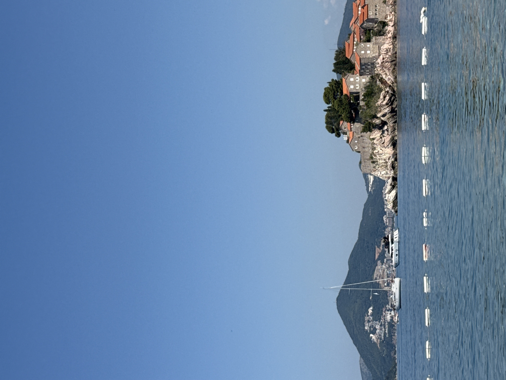 The image captures a coastal view in Budva, Montenegro, likely of Sveti Stefan island, under a clear, bright blue sky. In the foreground, calm blue water is marked by a series of white buoys. In the middle ground, a white sailboat with a tall mast is moored, with another, larger white yacht positioned to its right. Beyond the water, a rocky island is densely packed with stone buildings topped with red-tiled roofs, and numerous green trees. The island is connected to the mainland by a narrow causeway, though the causeway is not clearly visible. In the background, a large, verdant mountain rises steeply, dotted with numerous white buildings and structures. The overall scene conveys a sense of tranquil beauty and a popular tourist destination.