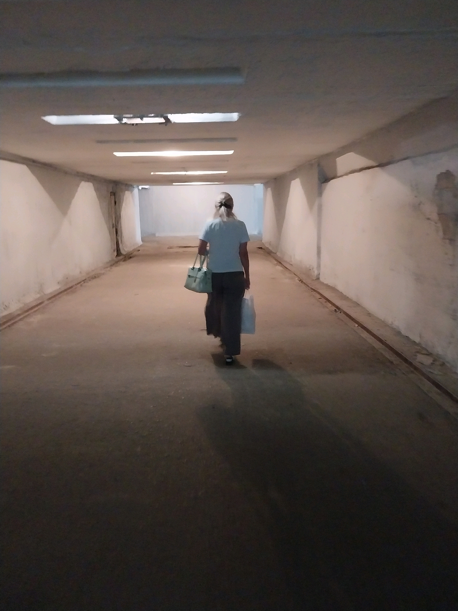A person is walking away from the camera down a dimly lit underground pedestrian underpass. The underpass has a concrete floor and walls, with fluorescent lights on the ceiling. The person is wearing a light blue t-shirt and dark pants and carrying two shopping bags, one light green and one white. The underpass appears to be in a state of disrepair, with cracked walls and debris on the floor. The lighting is poor, casting long shadows. The location is an underpass in the city of Dnipro, Ukraine.