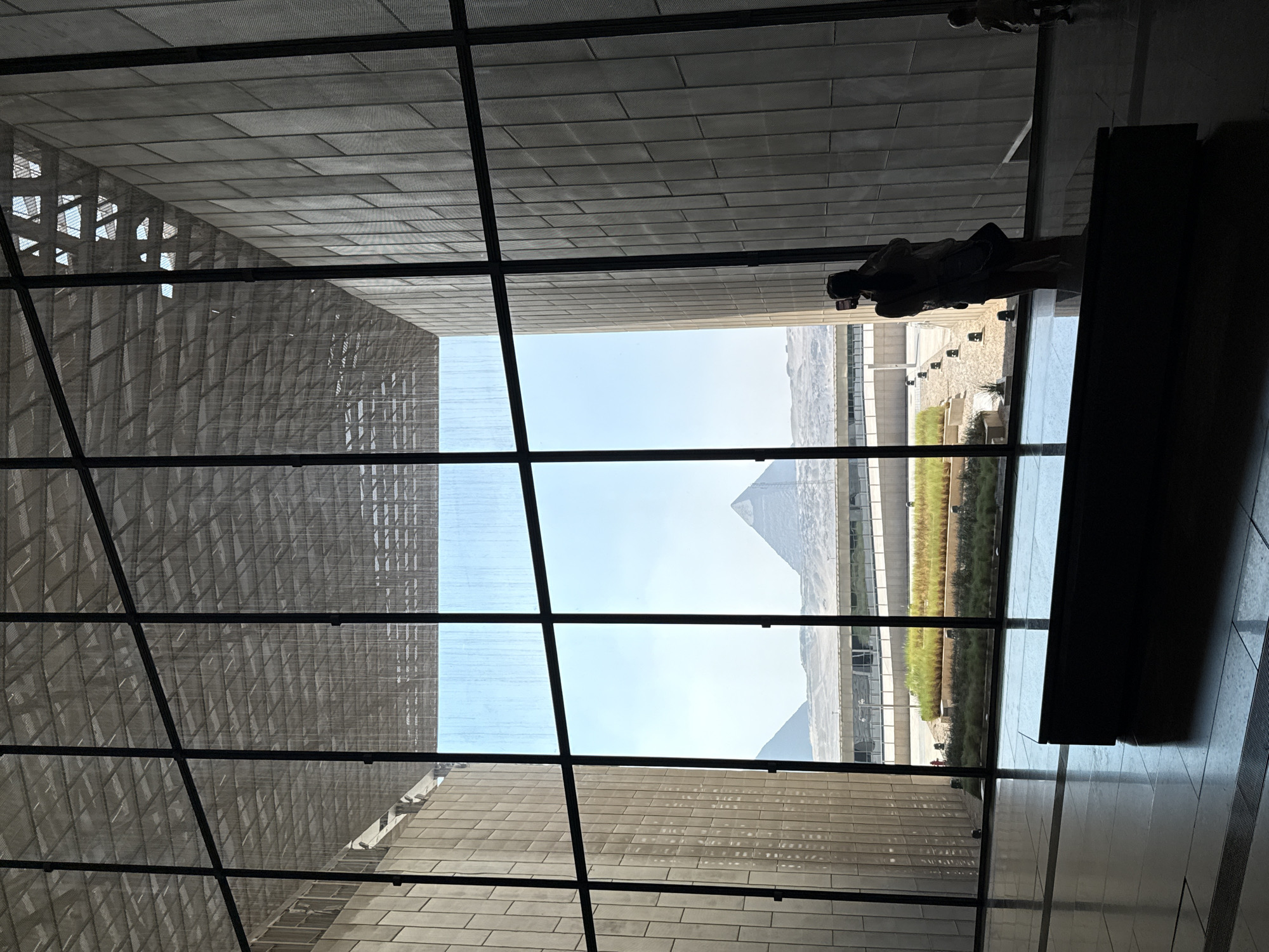 This is a wide-angle interior shot of a modern building with a large glass facade, offering a view of the Great Pyramids of Giza in Kafrat Nassar, Egypt. The interior features a high ceiling with a metallic grid structure and stone walls. A dark, rectangular bench is positioned in the foreground on the polished floor. Through the glass, the iconic pyramids are visible against a bright blue sky, with some modern structures and landscaping in between. In the middle ground, a person is silhouetted as they stand and take a photo with their phone. To the right, a child is walking away from the viewer, also silhouetted.