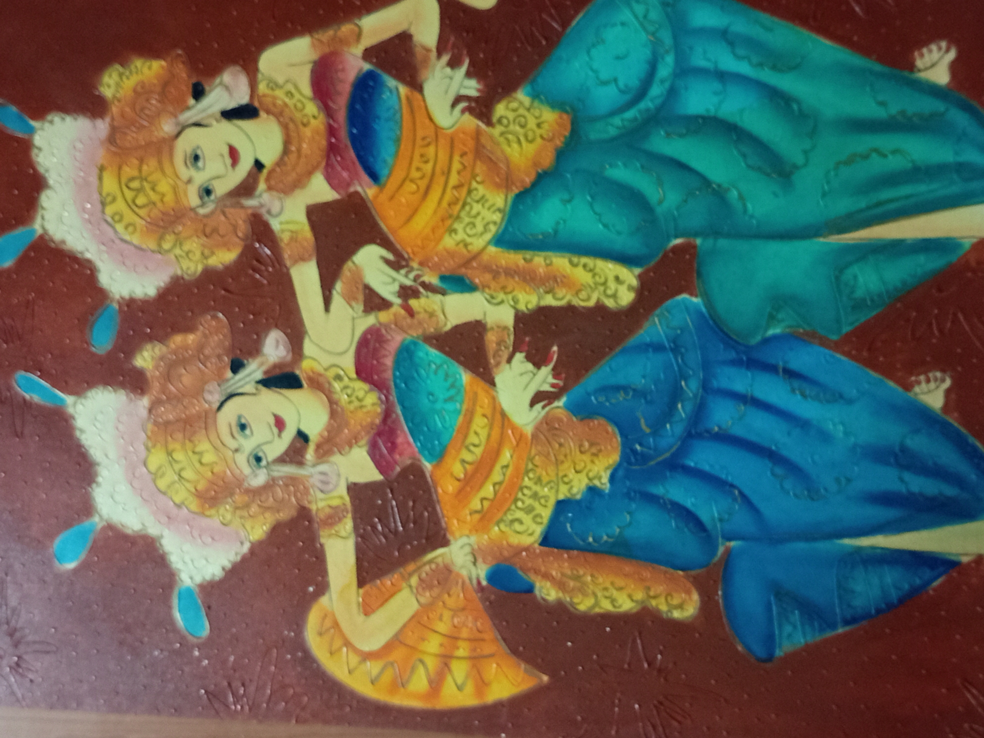 The image is a painting of two Balinese dancers in traditional attire. The dancers are depicted in mid-movement, with their arms outstretched and bodies in graceful poses. They are adorned with elaborate headdresses, ornate gold jewelry, and colorful, layered costumes in shades of red, blue, yellow, and green. The background is a textured, reddish-brown surface. The painting appears to be a decorative piece, possibly a souvenir or artwork from Bali, Indonesia. The specific location in Denpasar, Indonesia, is not evident from the image itself, as it depicts a general cultural scene rather than a specific landmark.