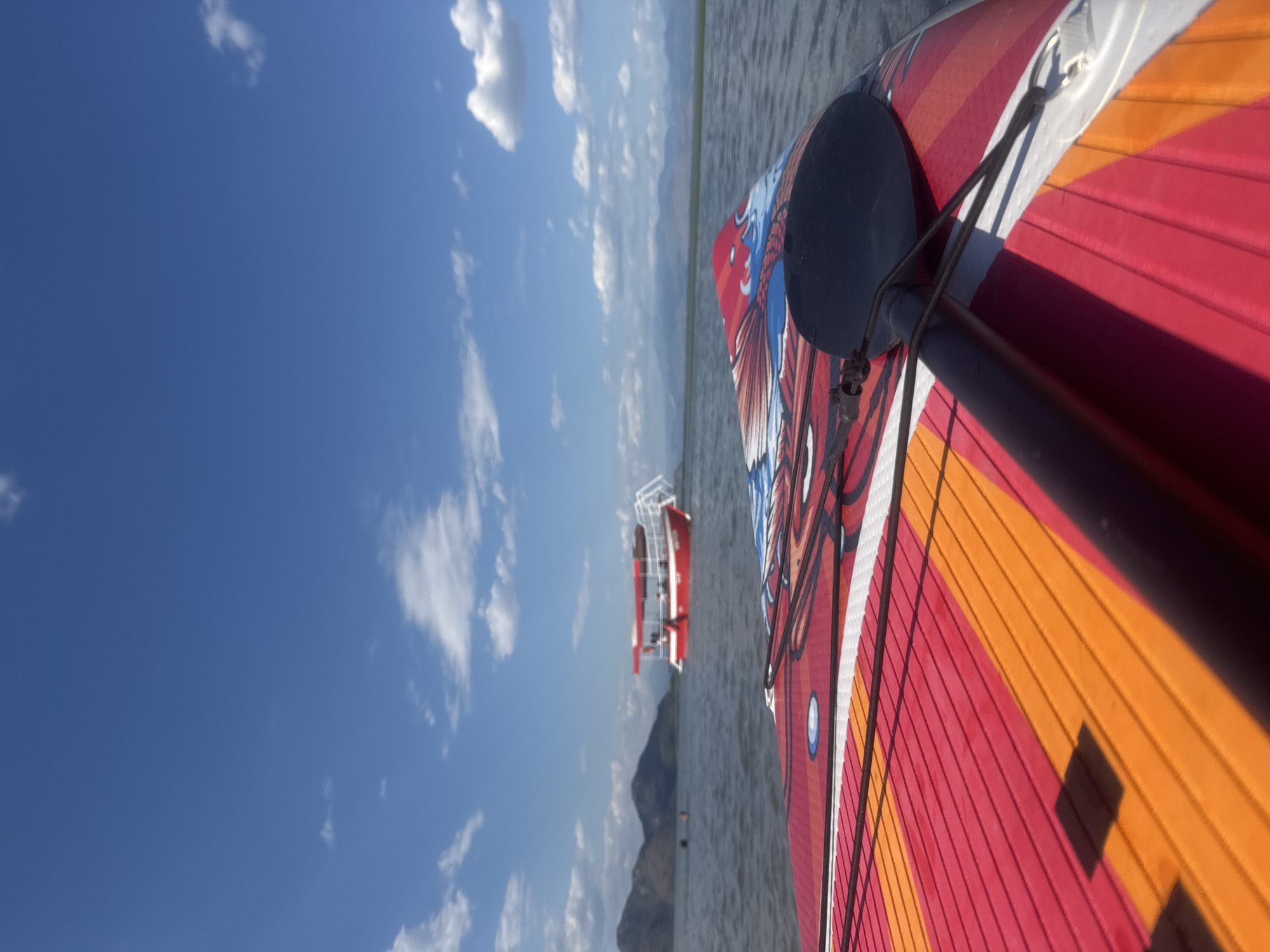 This is a first-person perspective, eye-level, close-up shot of a stand-up paddleboard on the water. The paddleboard is vibrant, with a red, orange, and white striped design featuring a blue and white graphic of a bird or dragon. A black paddle rests on the board, secured by black cords. In the background, a calm body of water, likely Skadar Lake, is visible. A red and white boat with a canopy and people on board is in the middle distance, and mountains form the horizon. The sky is a clear blue with scattered white clouds. The scene is set in the city of Seoca, Montenegro, specifically on Skadar Lake.