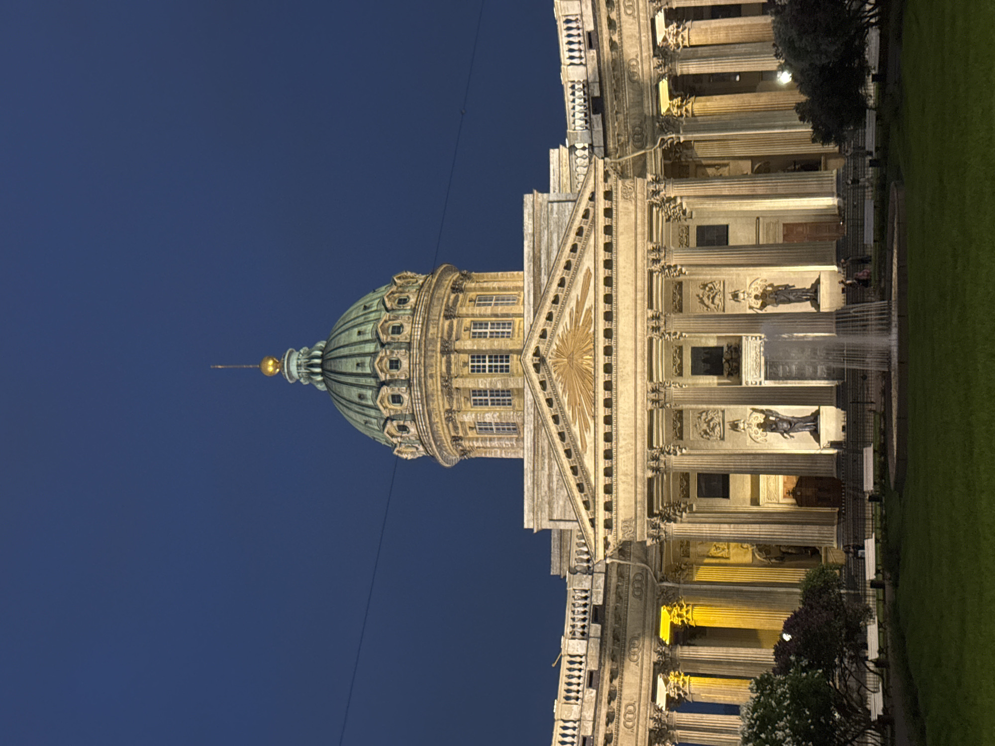 The image shows the Kazan Cathedral in Saint Petersburg, Russia, illuminated at night. The cathedral's dome dominates the left side of the frame, contrasting with the dark blue evening sky. The right side of the image depicts the cathedral's façade, featuring columns, sculptures, and architectural details. A fountain is visible in front of the building. A strip of green grass is located at the far-right of the frame.