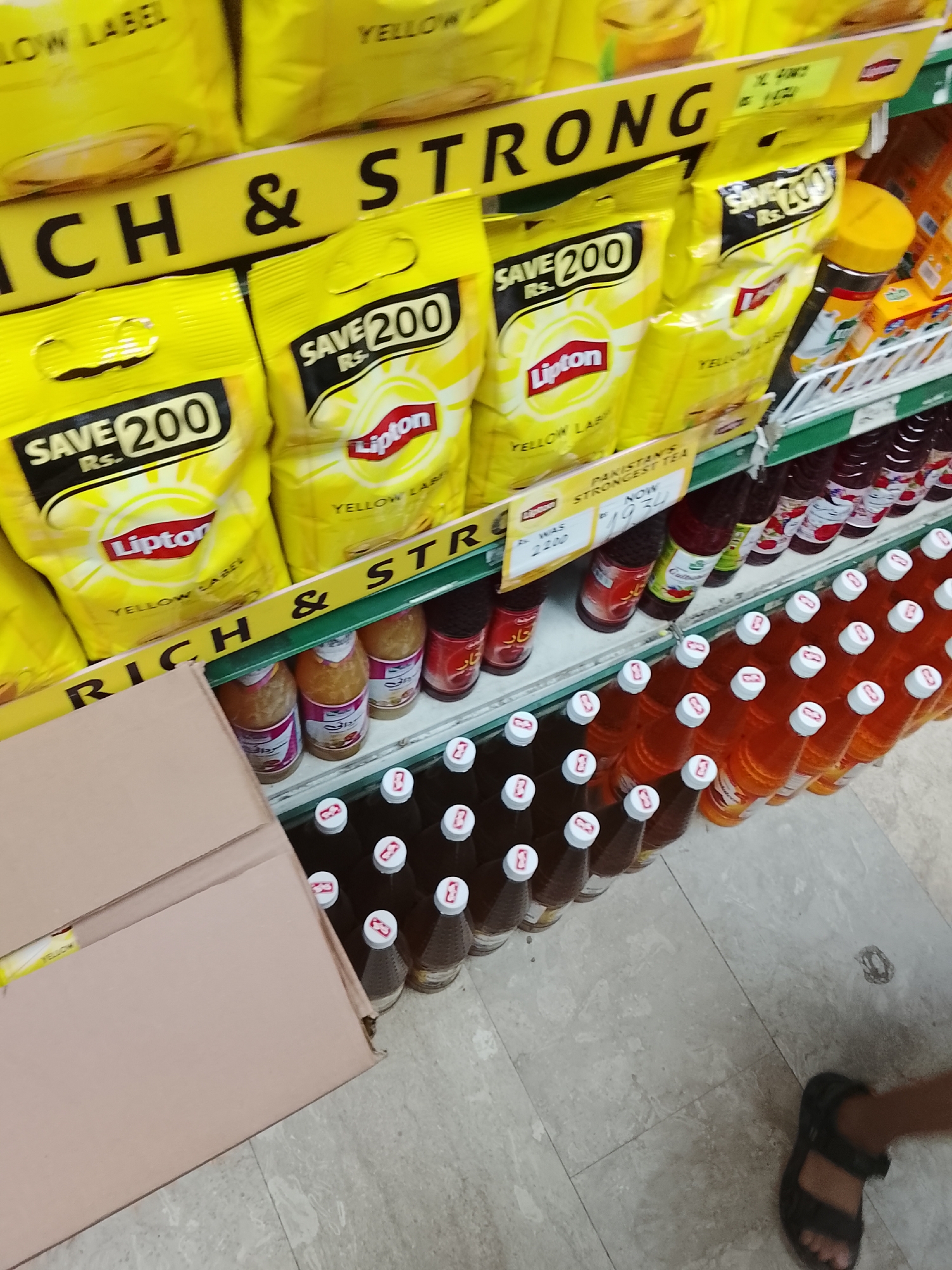 The image shows a retail display in a store, possibly located in Gulgasht Colony, Pakistan based on the product labeling ("Pakistan's Strongest Tea"). A cardboard box occupies the left foreground. On the shelves are several food and beverage items: Lipton Yellow Label tea bags with "SAVE 200 Rs." printed on the packaging, various bottled drinks (possibly juices or syrups) with red or orange colorings, and a few jars of a product labeled "Taar." A green shelf liner is partially visible. A person's foot, clad in a sandal, is visible in the lower right corner of the frame. The shelf is labeled "RICH & STRONG" with yellow signage. Promotional pricing for the Lipton product is visible: "Was 2200, Now 1934."