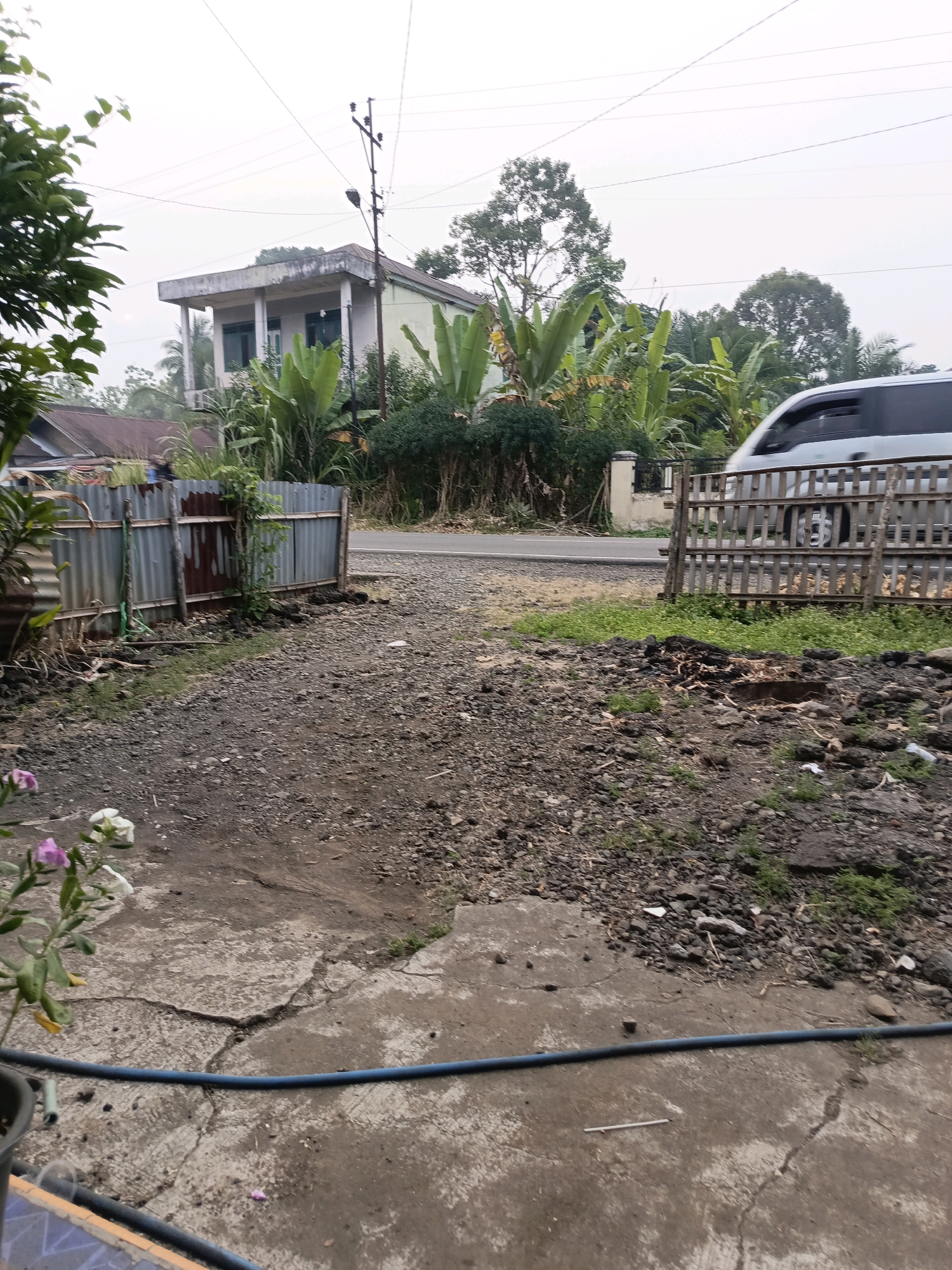 The image captures an outdoor scene, likely in Ulak Tanding, Indonesia, depicting a mixture of residential elements and natural vegetation. In the background, a two-story building with a light-colored facade and a dark roof is visible, surrounded by lush green banana trees and other foliage. A utility pole with power lines extends across the sky. To the right, a silver van is parked behind a wooden slatted fence. The foreground shows a rough, gravelly pathway leading towards a more paved, cracked concrete area. A small plant with pink and white flowers grows in the lower left corner. A blue hose lies across the concrete. The environment appears to be a somewhat rural or semi-urban area, with evidence of construction or land clearing in the middle ground, indicated by scattered debris and soil. The overall lighting suggests an overcast day.