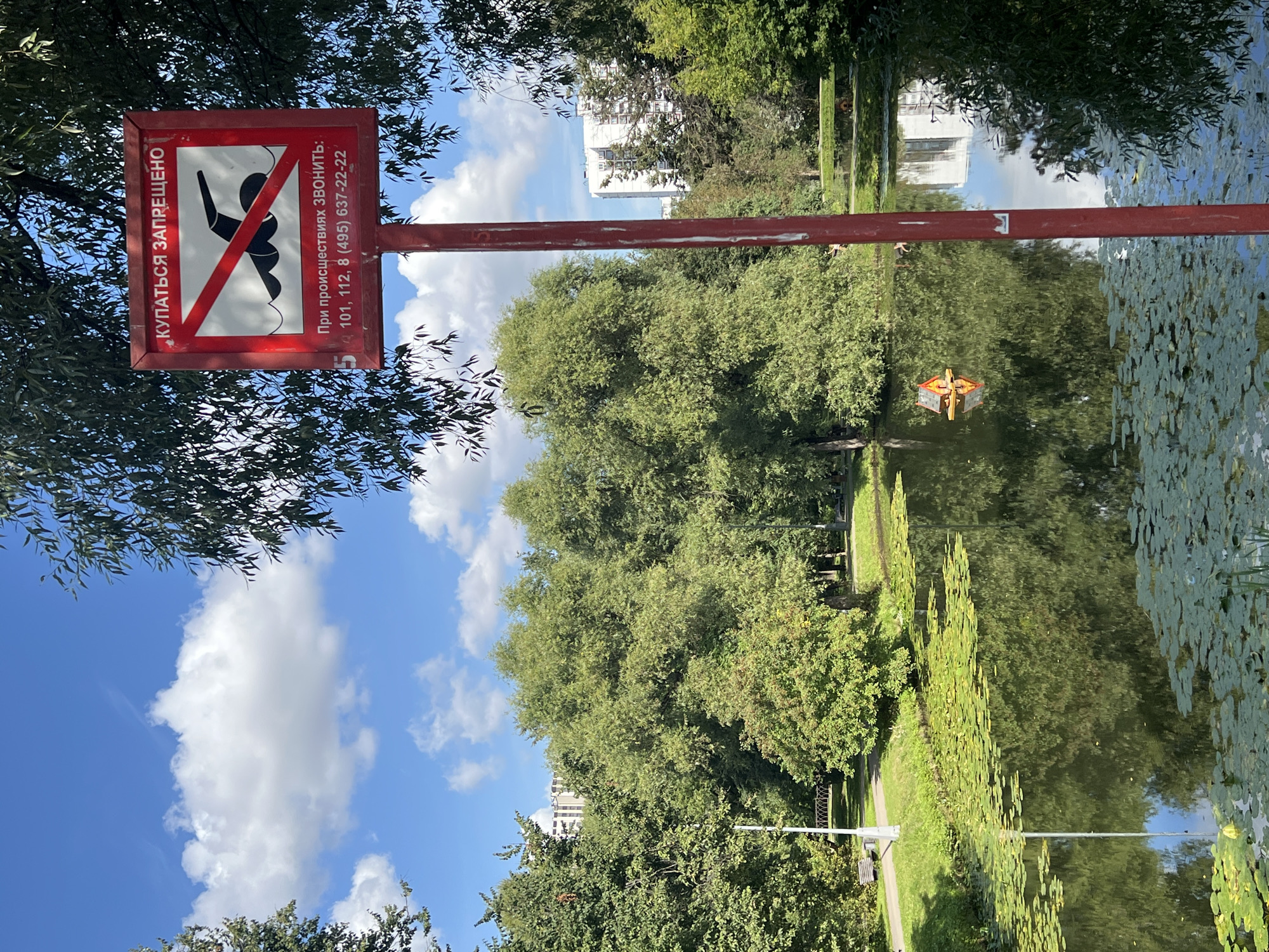 The image captures a scene in a park in Moscow, Russia, on a sunny day with scattered clouds. A prominent red sign on a pole reads "SWIMMING IS PROHIBITED" in Russian, featuring a graphic of a swimmer with a red diagonal line across it. Below this, it provides emergency contact numbers: "In case of emergencies CALL: 101, 112, 8 (495) 637-22-22". The sign is positioned near a tranquil pond covered with lily pads, reflecting the lush green trees and the blue sky. In the background, apartment buildings are visible, indicating an urban setting. The environment appears to be a public park with walking paths and benches, suggesting a recreational area. There are no people visible in the immediate vicinity of the pond or the sign.