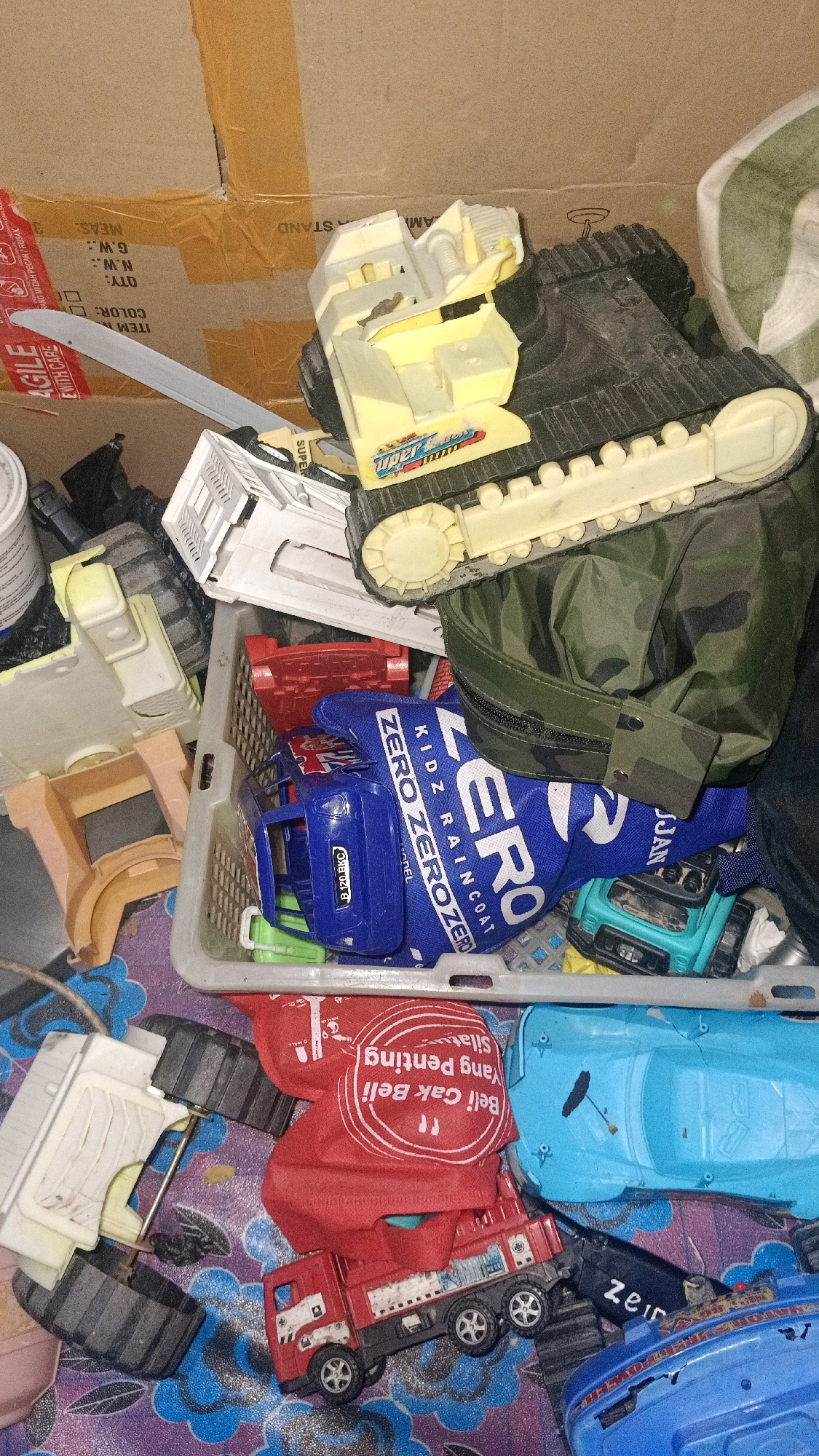 In the city of Central Jakarta, Indonesia, a collection of toys are piled in a cardboard box. The toys include a large black and yellow tank, a blue toy truck with "ZERO ZERO ZERO" written on it, a red toy truck, and a light blue toy car. There are also various plastic toy parts and a camouflage bag. The scene appears to be a disorganized storage area.