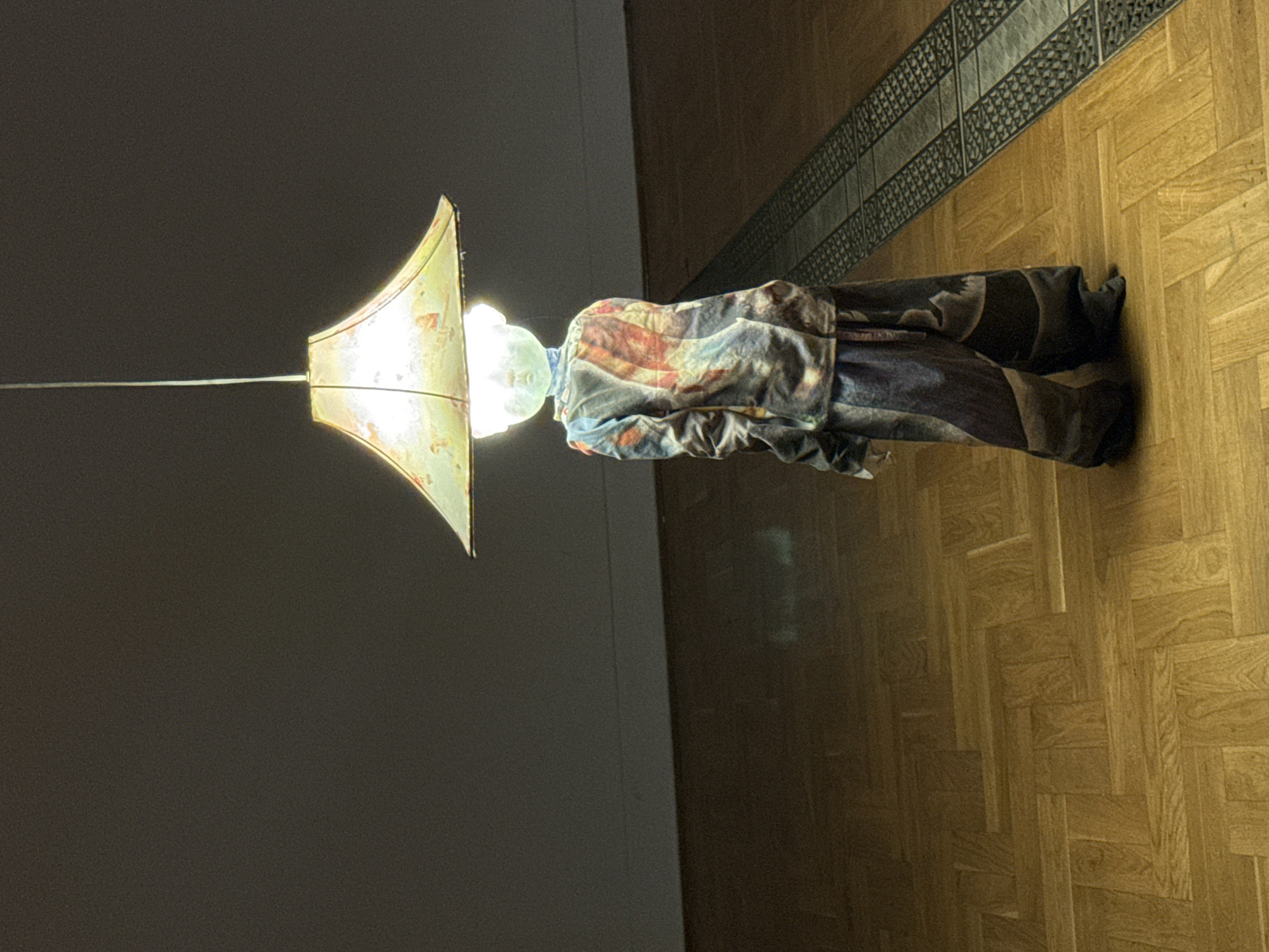 The image features a peculiar sculpture, seemingly depicting a headless figure wearing an intricately patterned garment and dark trousers. The figure is illuminated by a large, cone-shaped lampshade that is attached to a thin rod extending from the left side of the frame. The lampshade is translucent, allowing the light from within to cast a warm glow and highlight subtle designs on its surface. The figure's clothing is a riot of colors and patterns, with prominent strokes of red, blue, and white, resembling brushstrokes or abstract art.
The sculpture stands on a polished wooden floor, laid in a herringbone pattern, which reflects the ambient light. To the right of the figure, a decorative metal grate runs along the edge of the floor, bordering what appears to be a darker tiled area or wall. The background to the left of the sculpture is a plain, dark gray wall, creating a stark contrast with the brightly lit floor and the vibrant garment of the figure.
The lighting suggests an indoor setting, possibly a gallery or an exhibition space, due to the artistic presentation of the sculpture and the clean, well-maintained environment. There is no visible text within the image. The time of day and weather are not discernible from the scene. The overall impression is one of artistic expression and perhaps a touch of the surreal due to the headless nature of the figure and the unusual lampshade.