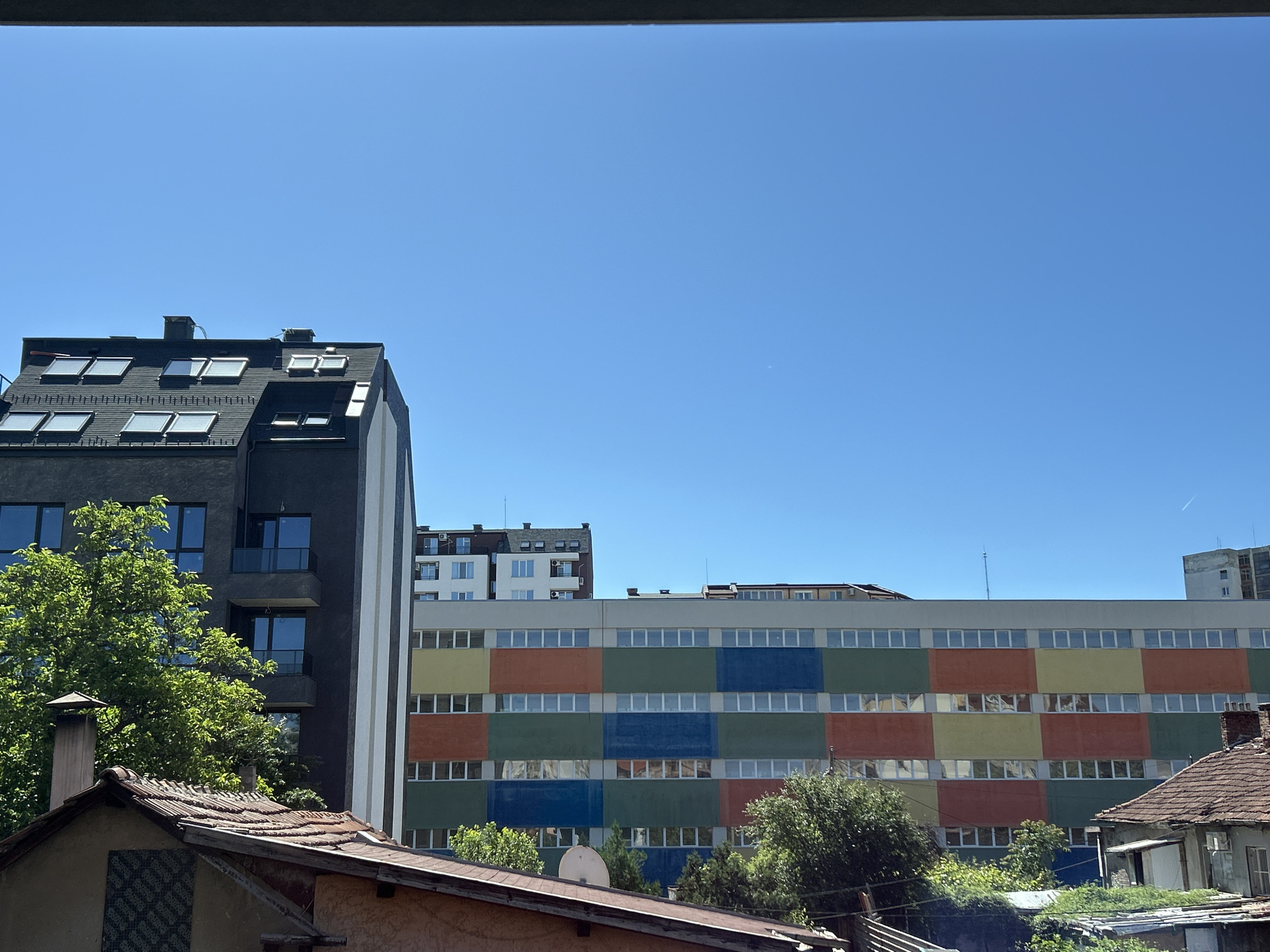 The image shows a view in Sofia, Bulgaria, featuring a mix of old and modern architecture under a clear blue sky. In the foreground are the rooftops of older, traditional buildings with terracotta tiles and weathered surfaces. Behind them rises a modern, dark-colored building with multiple floors and numerous windows. Adjacent to this newer structure is a large, rectangular building with a distinctive, colorful facade composed of alternating square blocks of orange, yellow, green, and blue. This building has a consistent row of windows on each floor. In the background, other buildings of varying heights are visible, contributing to the urban landscape. Vegetation, including trees, is interspersed throughout the scene, adding a touch of green to the urban setting. The scene is observed through a window frame.