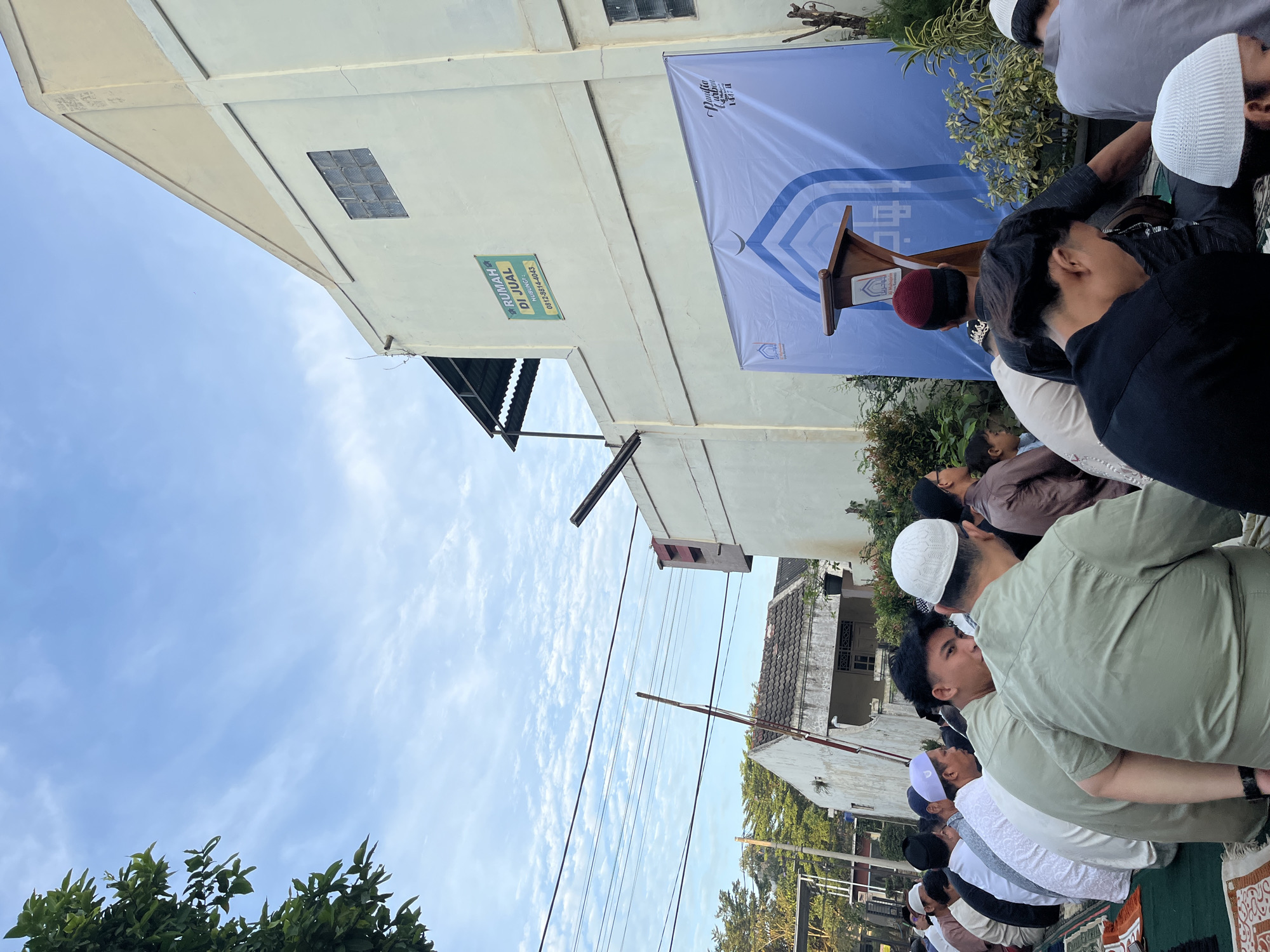 A gathering of Muslim men, most wearing white skullcaps, are praying outdoors on prayer mats. They are facing a makeshift podium with a blue banner behind it. The banner has text in Arabic. The background shows a building with a sign indicating "Rumah Di Jual". The overall location is determined to be in Sukabakti, Indonesia.