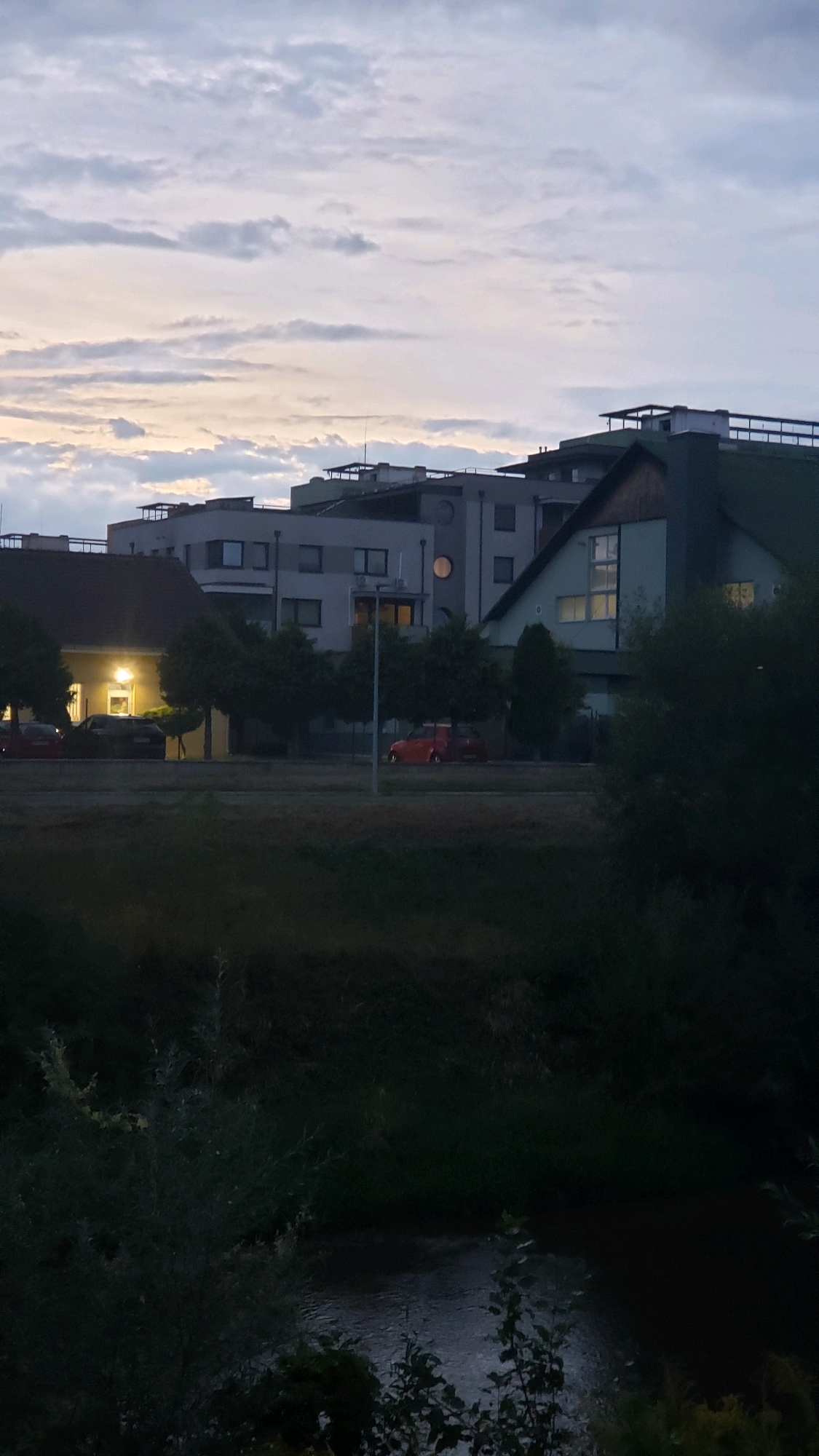 The image depicts a view of buildings and a street in Prešov, Slovakia, at dusk. In the foreground, there is dark foliage and a body of water, likely the Torysa River. Behind the foliage, a street is visible with several cars, including a red one parked or driving on the right side. On the left side of the street, a building with illuminated windows is present, possibly a residential or commercial property. Further in the background, multiple modern buildings with flat roofs and balconies are visible, along with a larger building with a green sloped roof. The sky is overcast with pale clouds, indicating the transition from day to night. There are no visible people in the image.