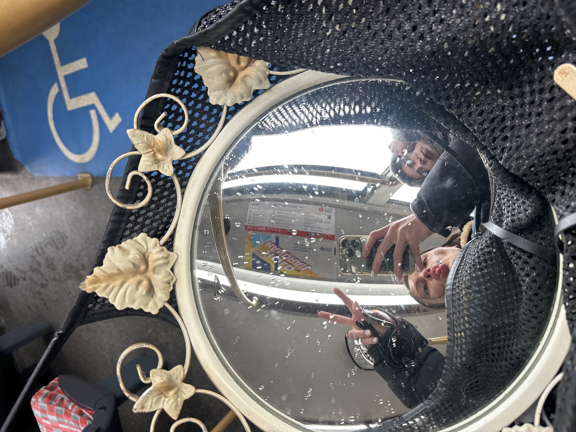 The photograph depicts a person taking a selfie in a round mirror. The mirror's frame is ornate, featuring floral designs. The reflection shows the person making a peace sign and holding a phone. The background visible through the mirror shows the interior of a London Underground train. A poster is partially visible with "Welcome" written on it. The environment is wet with water droplets on the surface of the mirror. A blue handicapped sign is visible in the background outside of the train.