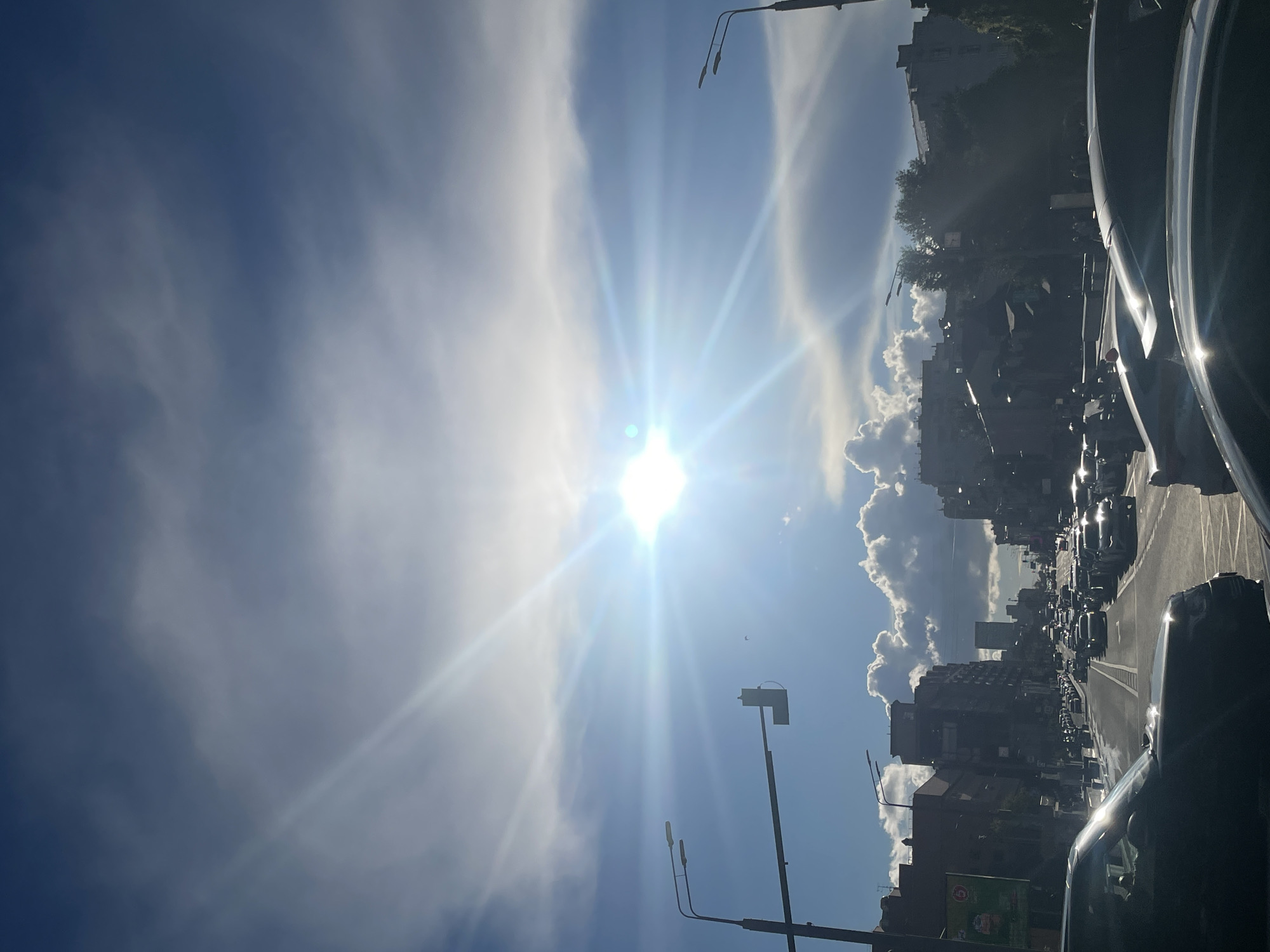The image depicts a street scene in Moscow, Russia, captured during the daytime. The sun is shining brightly, creating lens flares and casting rays across the sky. The sky is a vibrant blue, interspersed with wispy white clouds. In the foreground, the side of a dark-colored car is visible, reflecting the sunlight. The road ahead is lined with buildings of varying heights, some appearing to be residential apartments and others office buildings. A significant traffic jam is evident, with numerous cars stretching down the street. Streetlights are also visible, positioned along the roadside. The overall impression is of a busy urban environment on a sunny day, with the sun's intensity being a prominent feature of the scene. There is no visible text in the image.