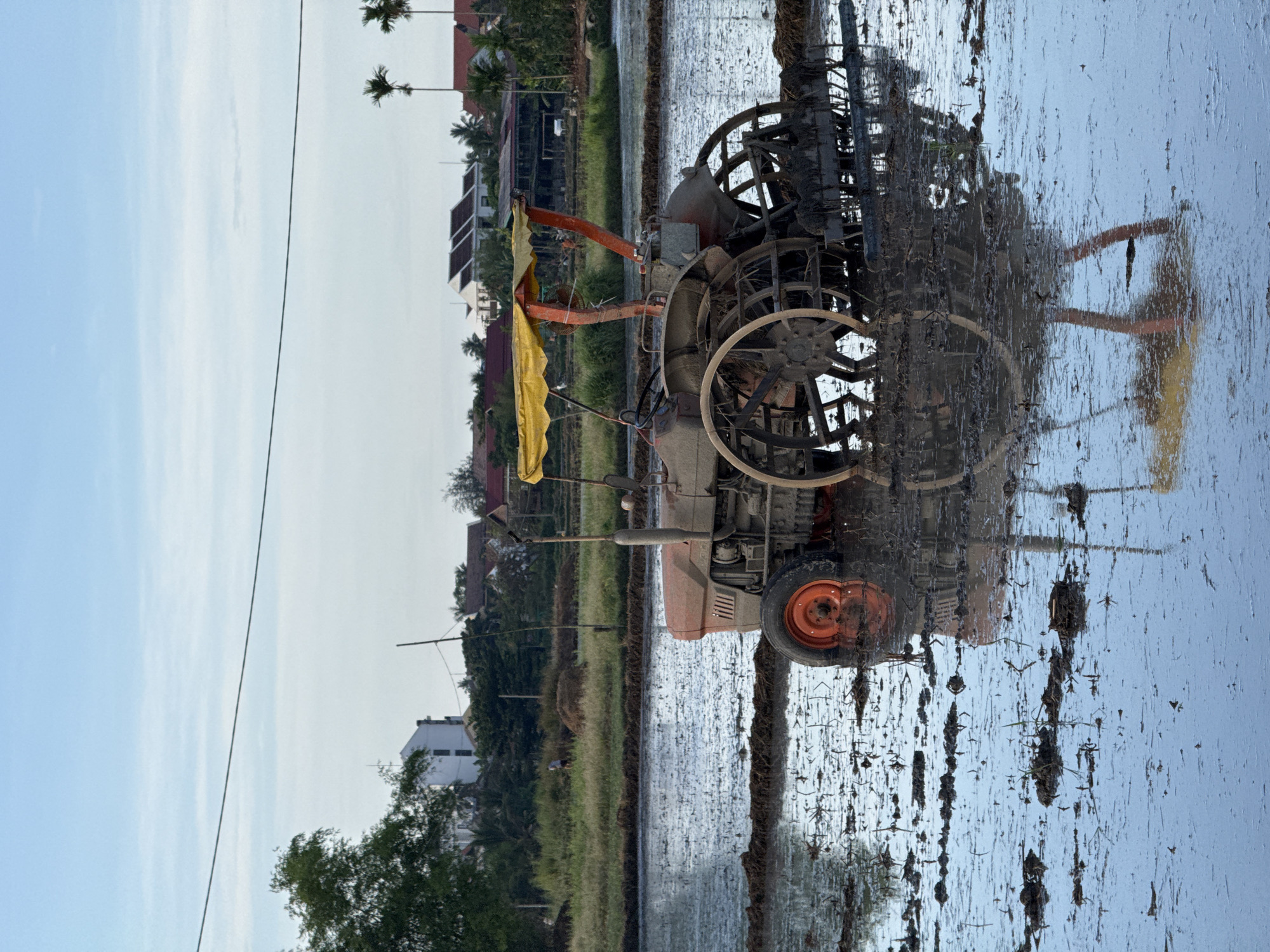 This image presents a rural landscape within the city of Đà Nẵng, Vietnam. The central subject is an orange tractor, partially submerged in a flooded rice paddy. The tractor features an orange front left rubber tire and large, mud-encrusted metal cage wheels at the rear, with a yellow canopy mounted above the operator's area. A muddy agricultural implement is attached to the tractor's rear. Clear reflections of the tractor and the pale blue sky are cast upon the shallow, muddy water, which contains minimal organic debris. In the middle distance, a band of green vegetation separates the paddy from a residential area. A solitary person is visible on the left, near a mound of hay or earth. The background displays a mix of white multi-story buildings and traditional houses with dark roofs, interspersed with palm trees. A thin black power line stretches horizontally across the upper frame. The sky is light blue with sparse, faint cloud formations.