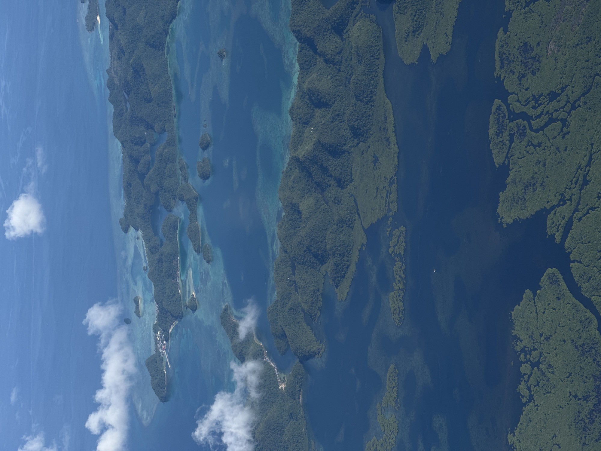 The image shows an aerial view of a coastal area with numerous islands and waterways, likely located near Del Carmen, Philippines. Dense vegetation, primarily consisting of mangrove forests, covers many of the islands. The water varies in color, ranging from clear turquoise near the coast to deeper blues in the open sea. Some structures and small settlements are visible on a few of the islands, suggesting human habitation. A few white clouds are visible, some near the horizon and others dotted in the sky.