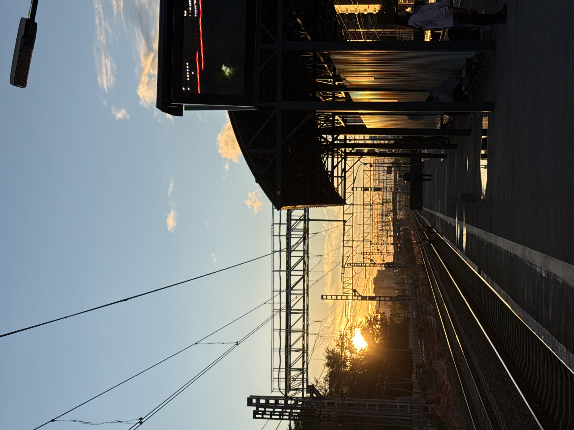 The image depicts a train station platform at sunset. The sun, low in the sky, casts a warm, golden glow through the clouds and illuminates the corrugated metal roof of the station's overhang. The tracks of a railway stretch into the distance, reflecting the light. The scene is viewed from a slightly elevated perspective, looking down the platform.
In the foreground, a dark, rectangular object, possibly a light fixture or a sign, hangs from a pole on the left side of the frame. Above the platform, a complex network of overhead wires and support structures for the electrical system is visible against the clear, blue sky. The sky is dotted with wispy, pale clouds that are catching the last rays of sunlight.
Several people are present on the platform. One person, dressed in a light-colored jacket, stands near the edge of the platform on the right. Further down the platform, another person is seated, and other figures are silhouetted against the light, suggesting movement and activity. The overall mood is serene and tranquil, amplified by the beautiful sunset.
The location is suggested to be Moscow, Russia. The presence of an overhead railway system and the architectural style of the station are consistent with such a location. The time of day is clearly sunset, indicated by the low angle of the sun and the warm, diffused light. The weather appears clear and pleasant. There is no discernible text in the image.